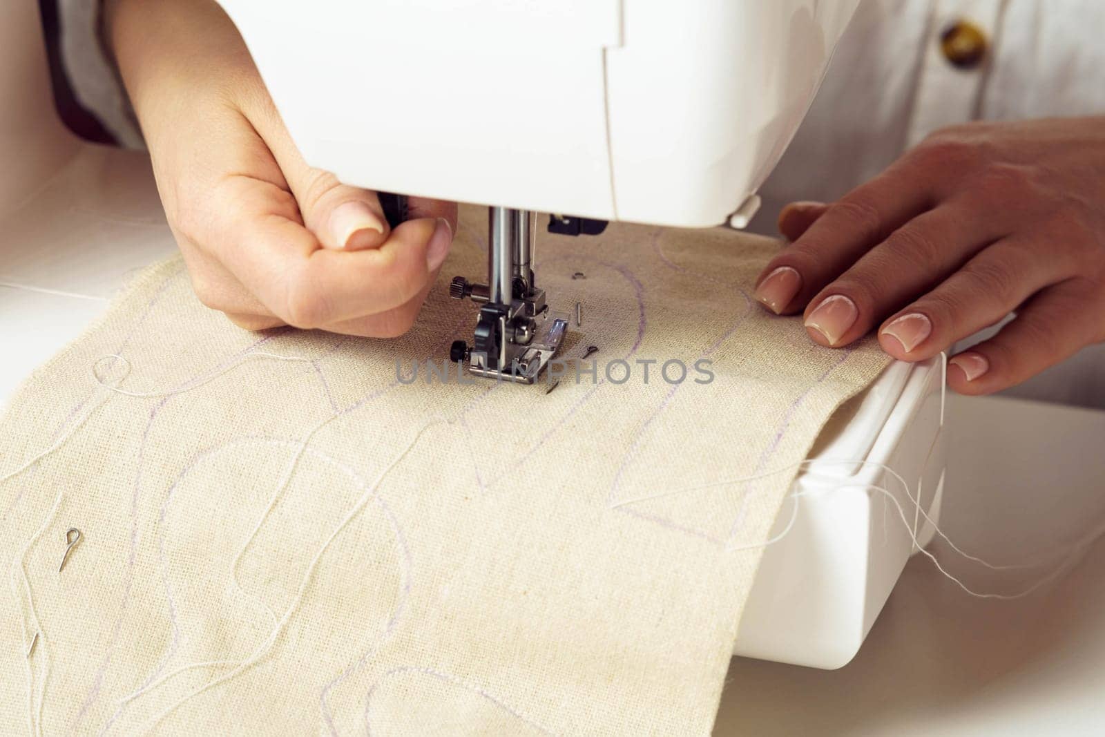 Cropped woman dressmaker hands using sewing machine, putting white soft fabric with stitch contour on needle plate under presser foot. Clothes production in workshop. Creative small business. Closeup