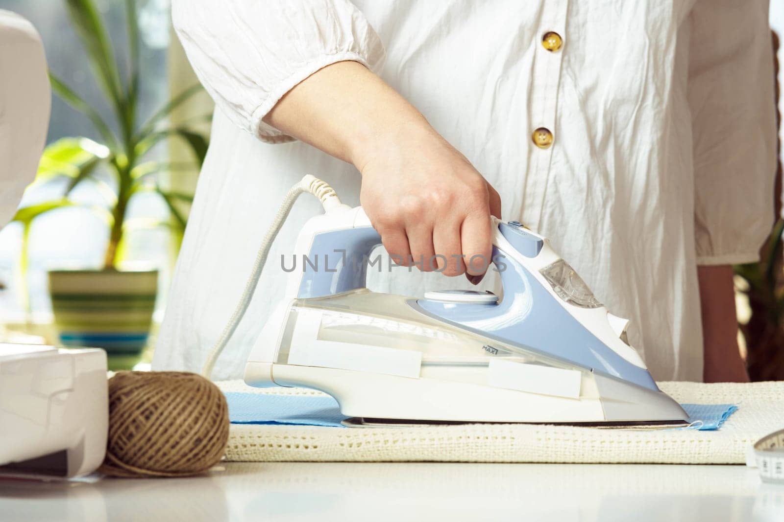 Cropped woman wearing white shirt steam iron blue wrinkled textile on table neat thread yarn. Dressmaker prepare fabric for sewing, manufacturing work indoors. Small business or hobby. Close up