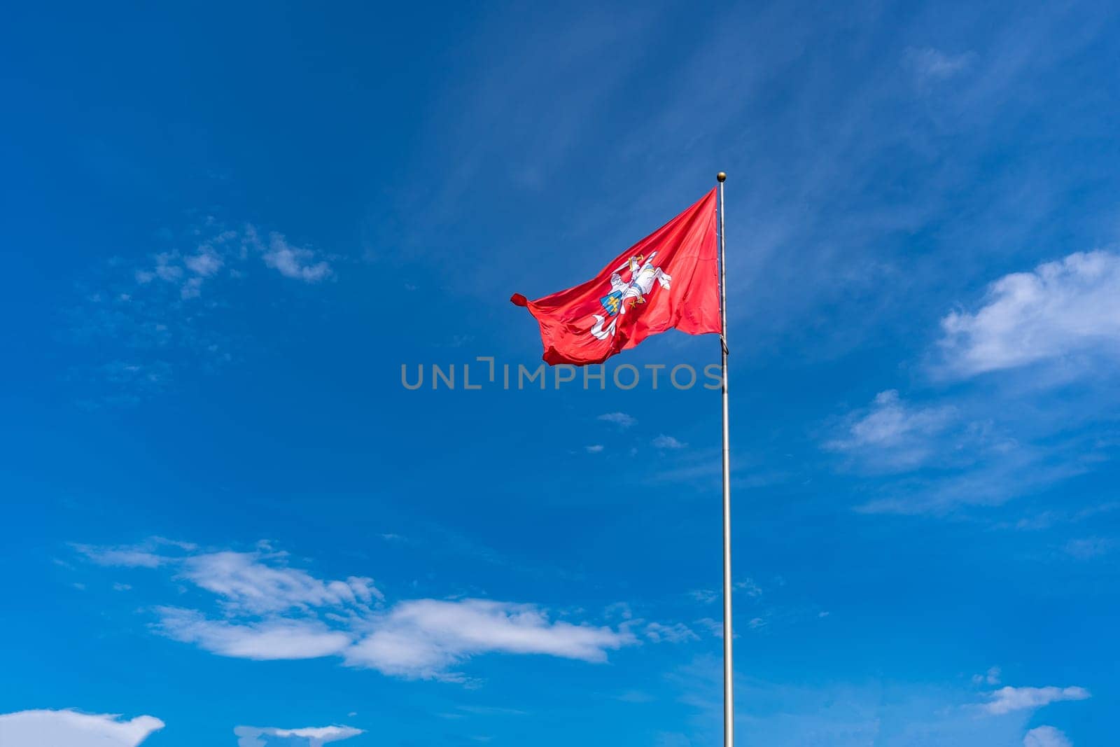 The historic flag of Lithuania, a rider with a sword on a white horse on a red background flutters against a bright blue sky with white clouds.