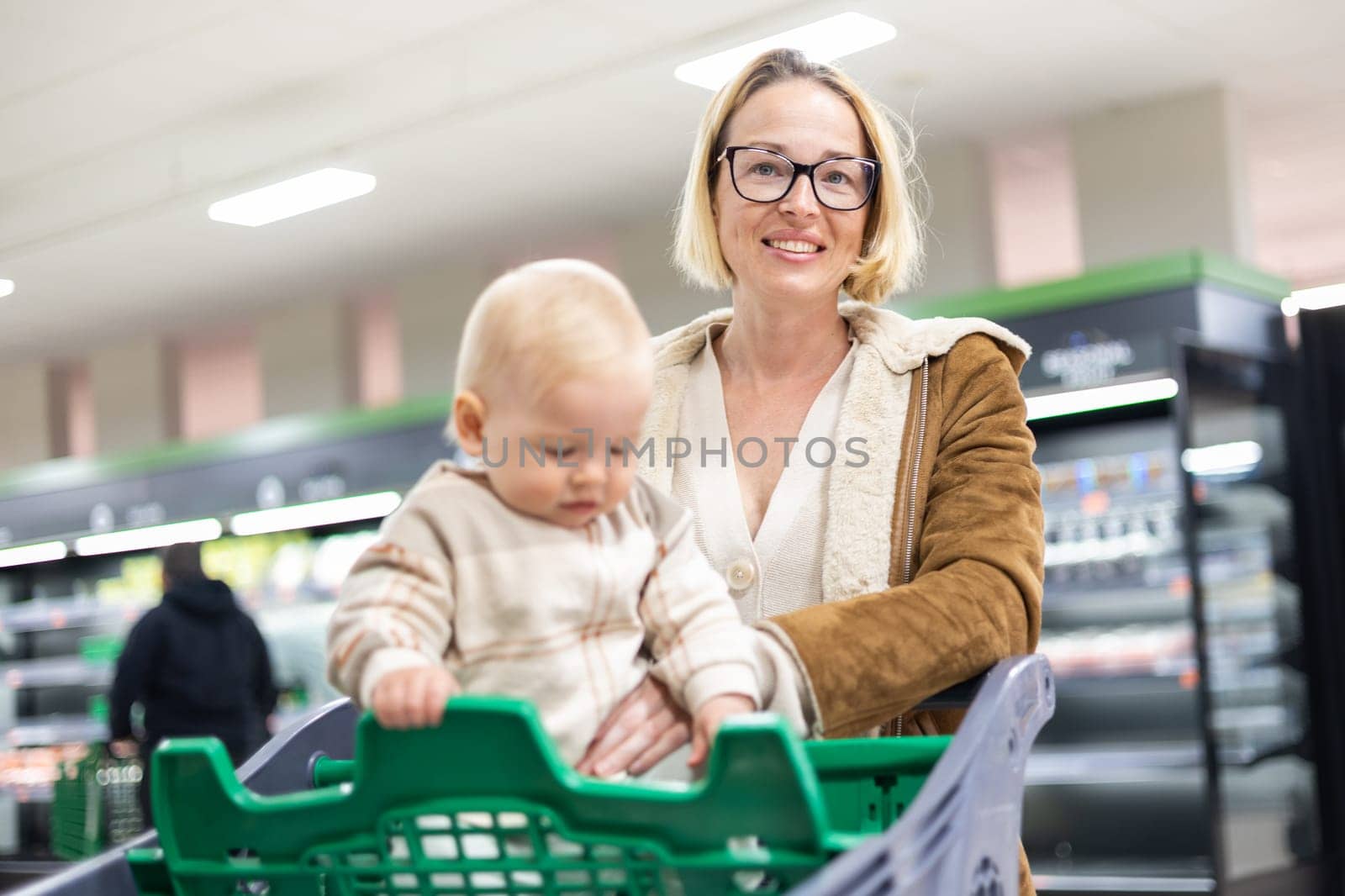 Caucasian mother shopping with her infant baby boy child choosing products in department of supermarket grocery store