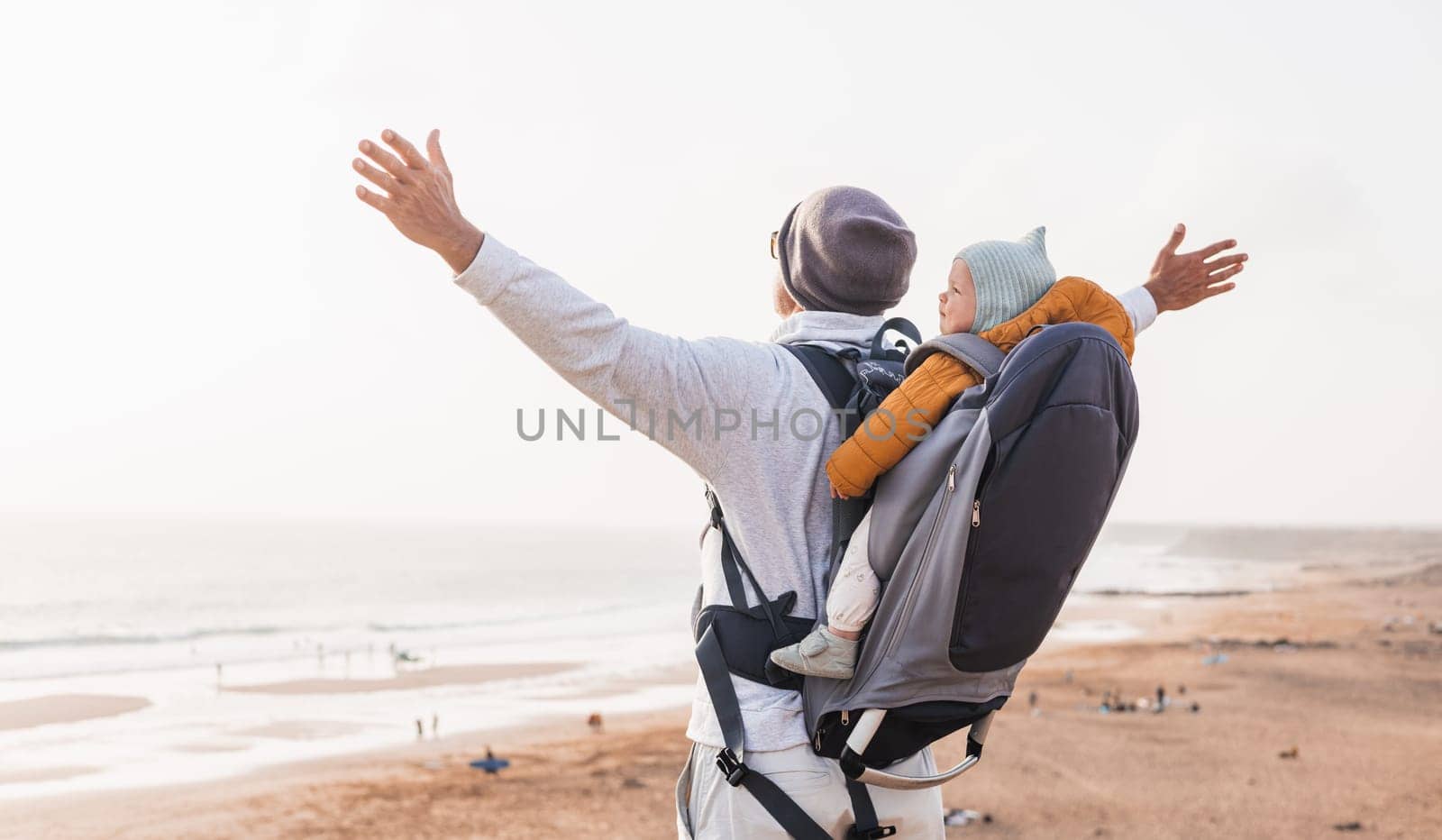 Young father rising hands to the sky while enjoying pure nature carrying his infant baby boy son in backpack on windy sandy beach. Family travel concept