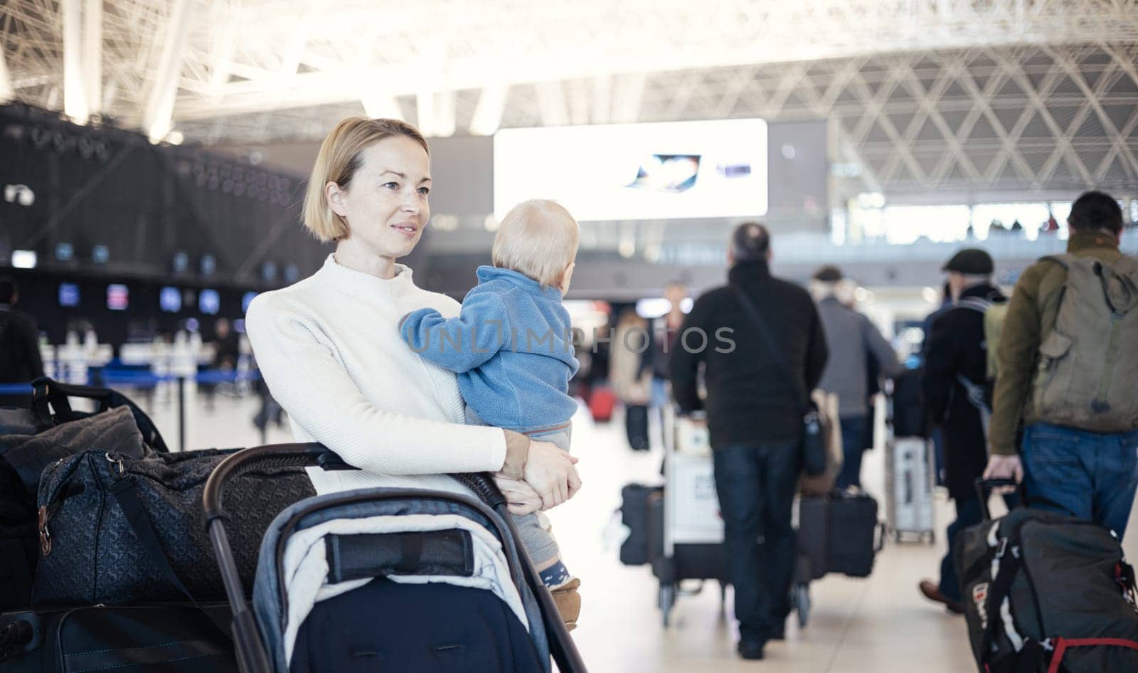 Motherat travelling with his infant baby boy child, walking, pushing baby stroller and luggage cart at airport terminal station. Travel with child concept