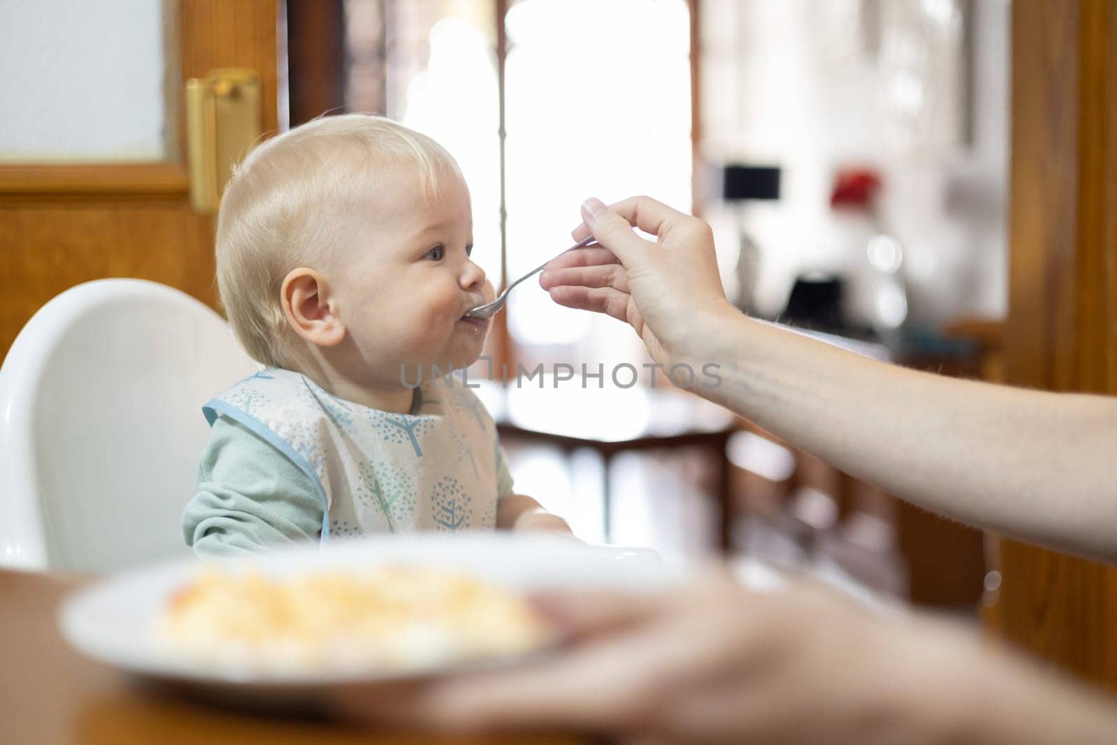 Mother spoon feeding her infant baby boy child sitting in high chair at the dining table in kitchen at home.