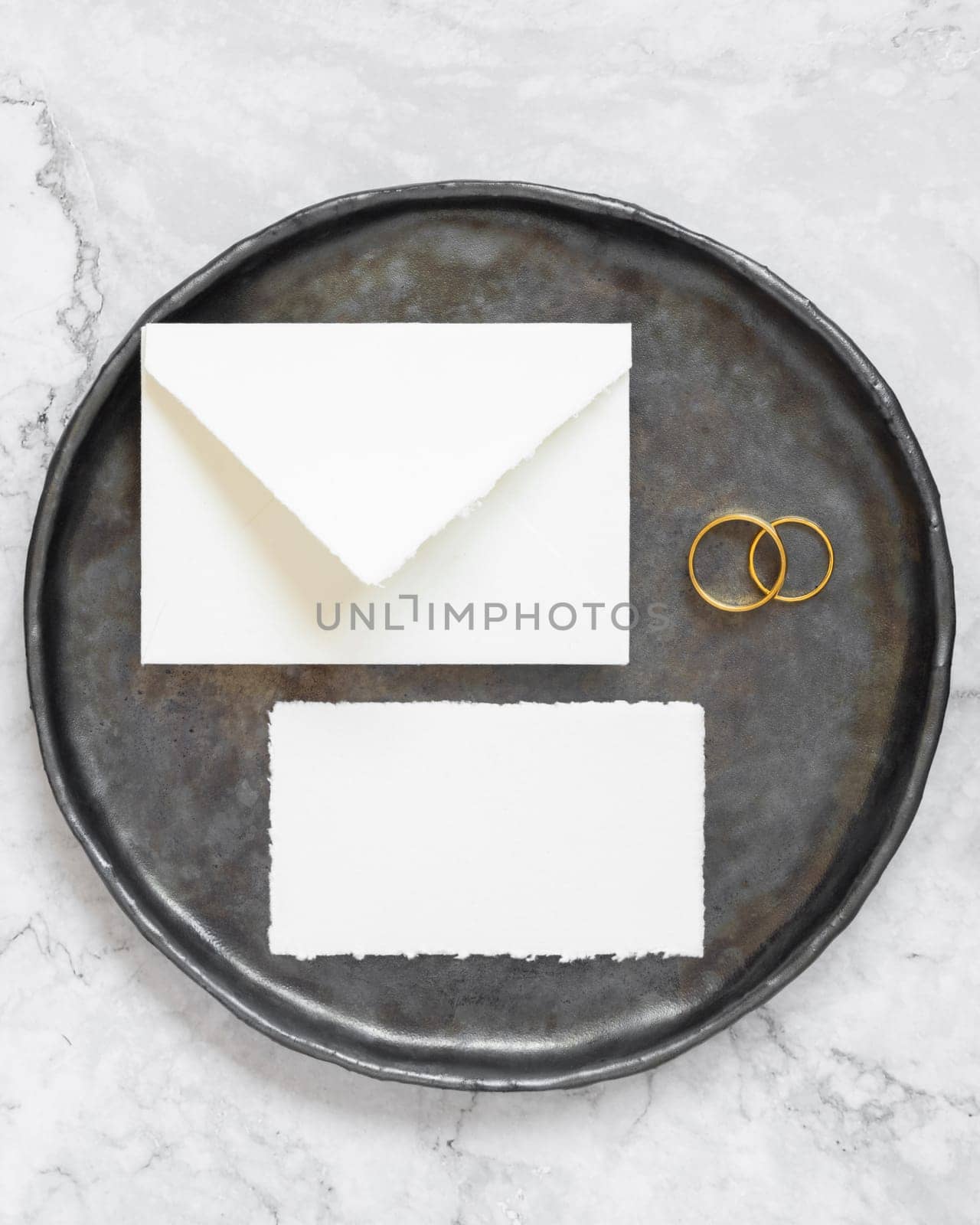 Black plate with a blank card, envelope and wto wedding rings top view, mockup. Elegant flat lay with horizontal greeting or Invitation paper card and envelope, wedding or holiday stationery