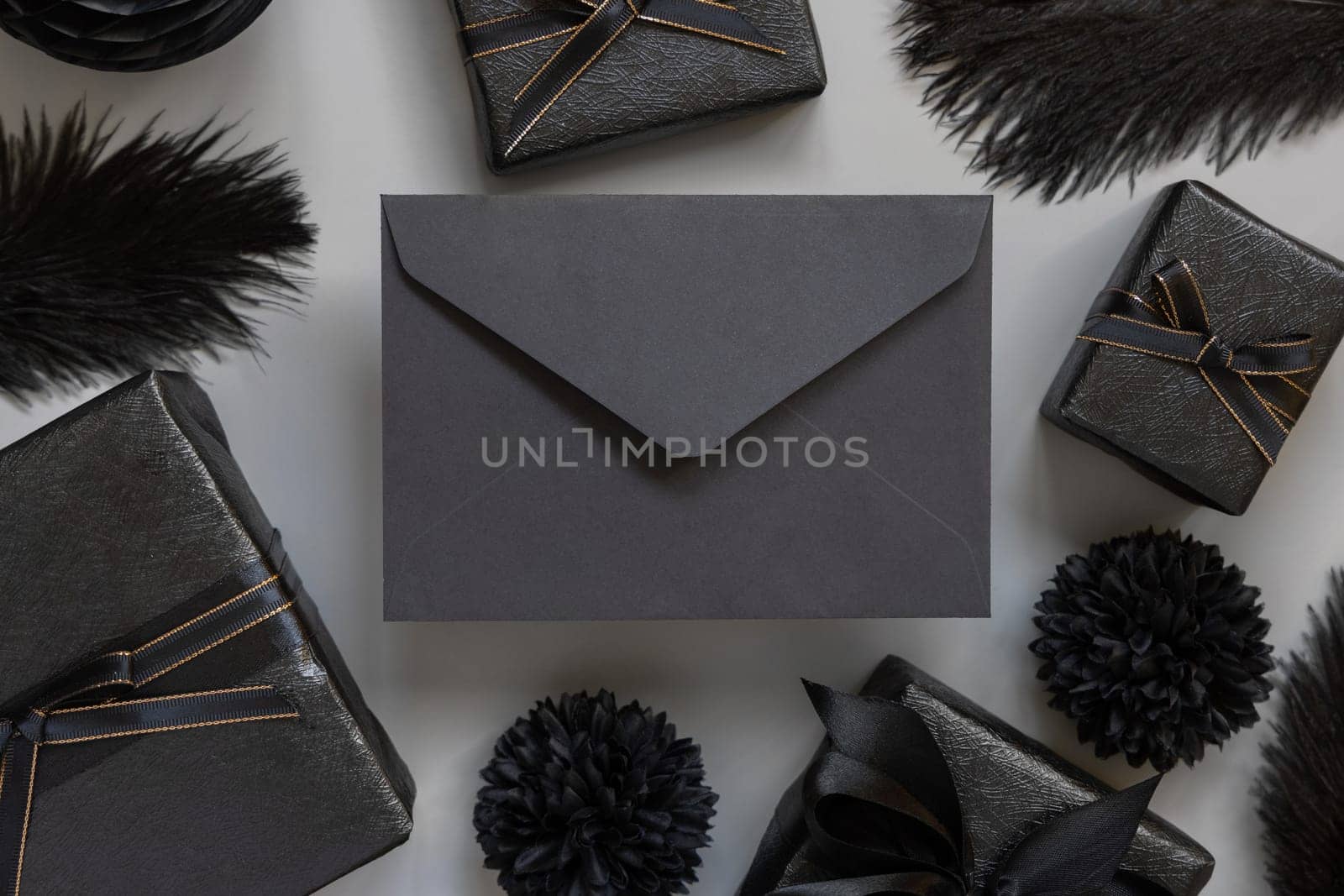 Black envelope between Wrapped Gift Boxes, black paper flowers and feathers, mockup, copy space. Dark composition with horizontal paper card for Birthday, Anniversary or black party present