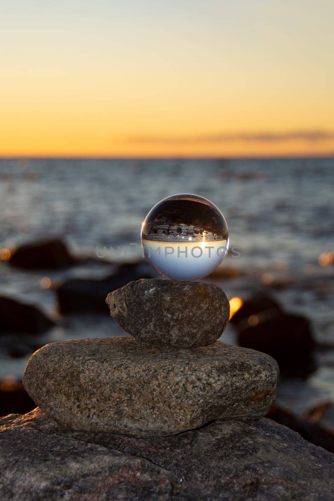 Glass ball lies on the top stone of stacked stones, at sunset on the beach, the sea and the setting sun are reflected in the ball