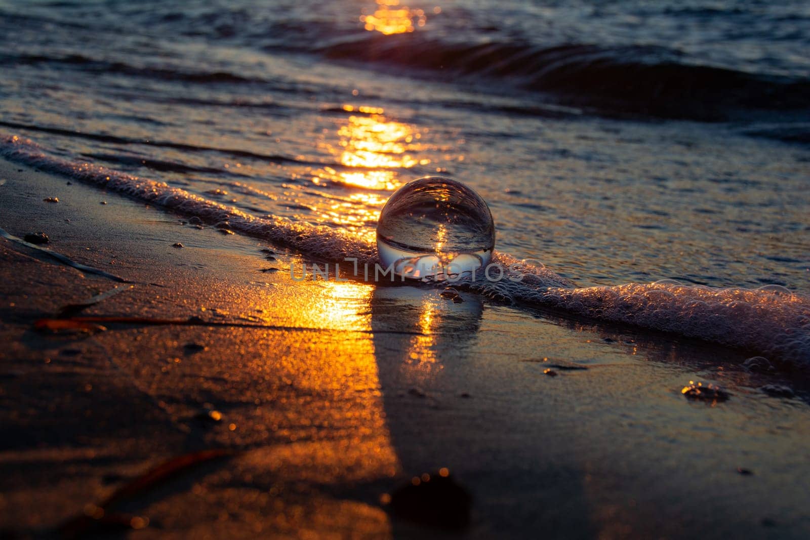 A glass ball lies in the waves on the sandy beach, the sea and the setting sun are reflected in the ball