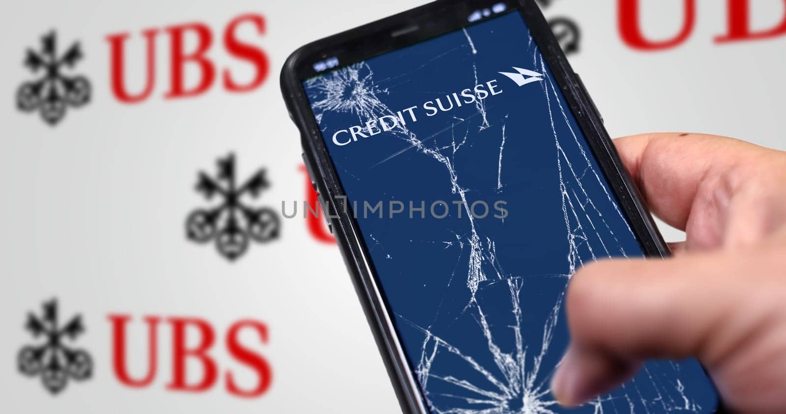 New York, US, March 2023: Hand holding a phone with Credit Suisse logo on cracked screen. UBS logo blurred on white background. UBS bought Credit Suisse for 3B CHF in stock. Illustrative editorial