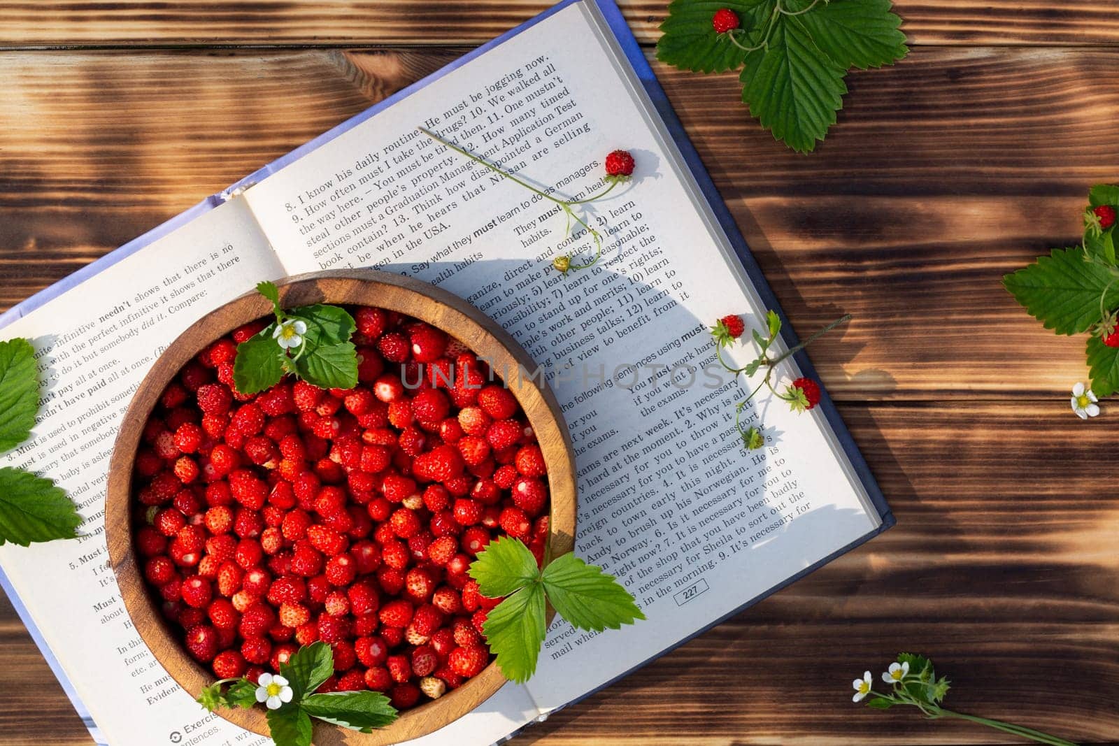 A wooden bowl of red ripe wild strawberries and flowers with a book on an old wooden surface. Top view. Copy space.