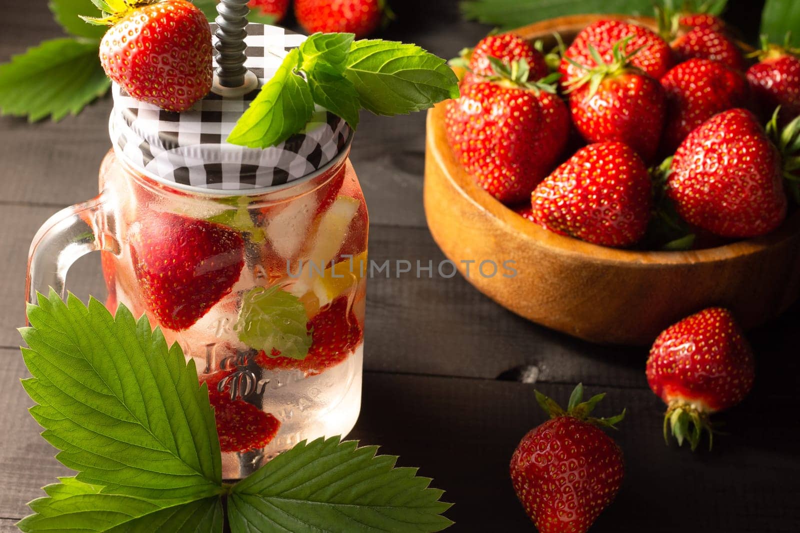 Summer cold drink with mint, ice, lemon and strawberries and a wooden bowl of fresh strawberries on a black wooden surface.