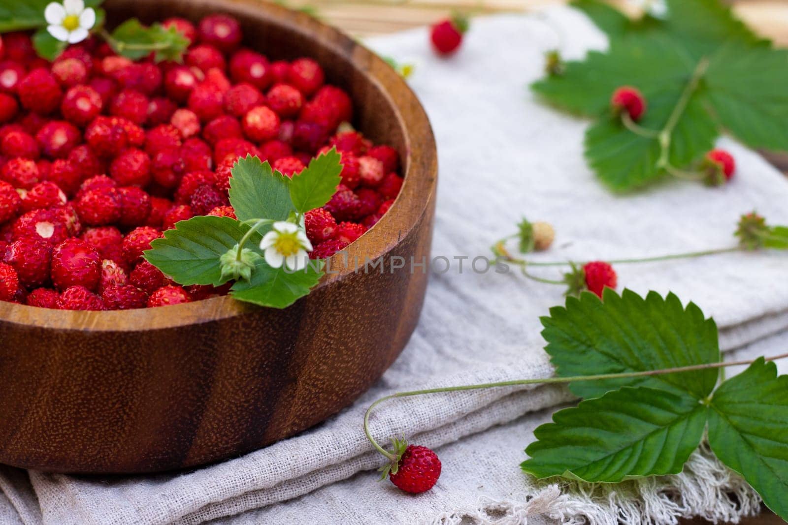A wooden bowl of red ripe wild strawberries and flowers on an old wooden surface. Copy space.