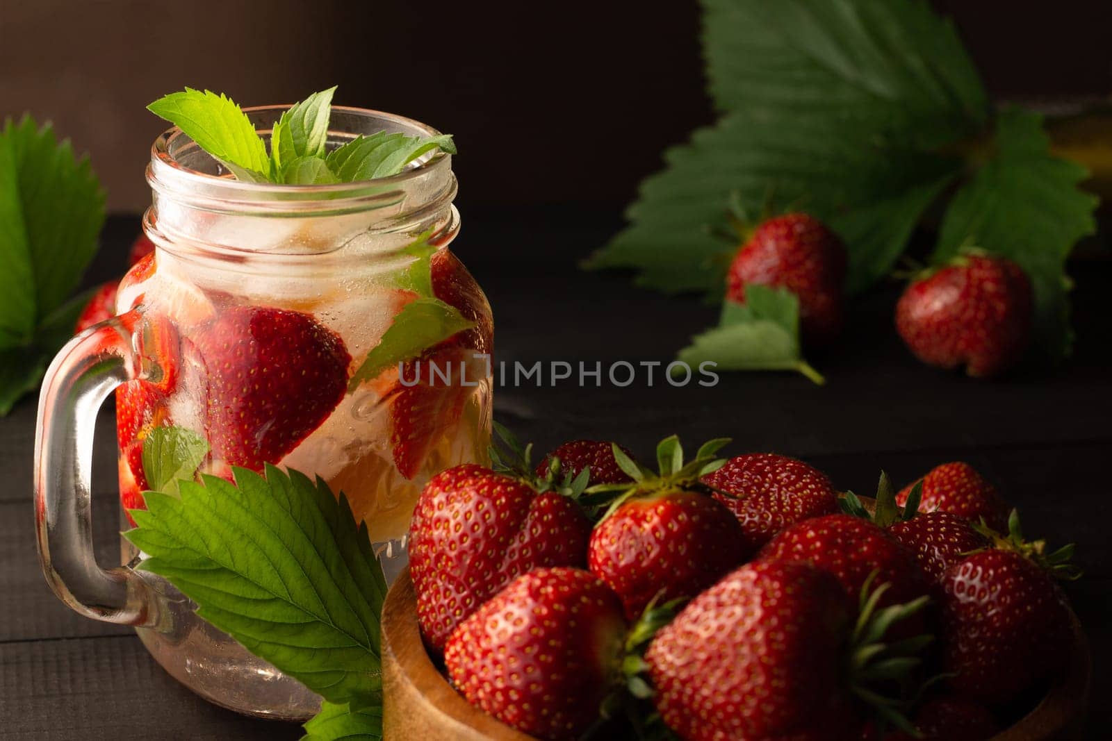 Summer cold drink with mint, ice, lemon and strawberries and a wooden bowl of fresh strawberries on a black wooden surface. Low key