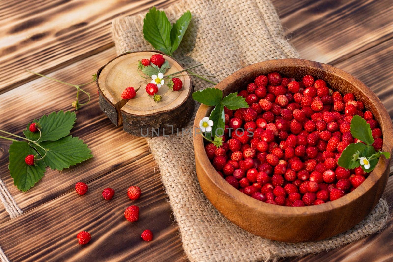 A wooden bowl of red ripe wild strawberries and flowers on an old wooden surface.