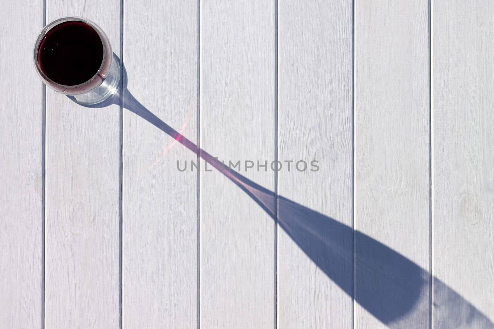 A glass of red wine and its long shadow on a white plank background. Top view, copy space