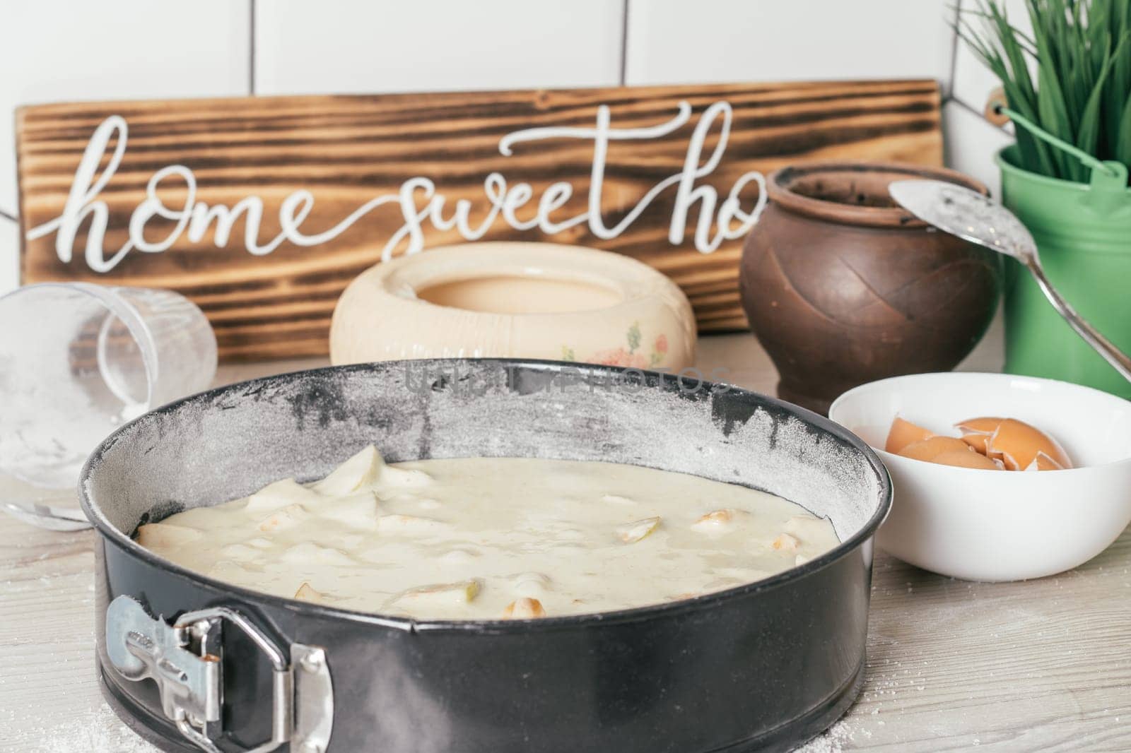 Baking dish with raw apple pie dough stands on kitchen counter next to ingredients, cooking utencils and wooden sign Home sweet home