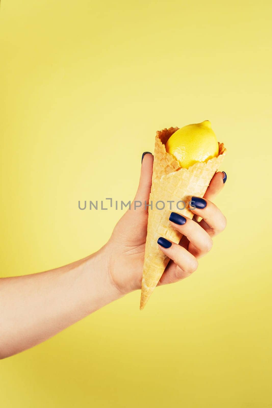 Female hand with blue nails holding waffle ice cream cone filled with fresh ripe juicy lemon isolated on yellow. Creative art concept. Food minimal colorful concept. Vertical. Copy space. Mock up.