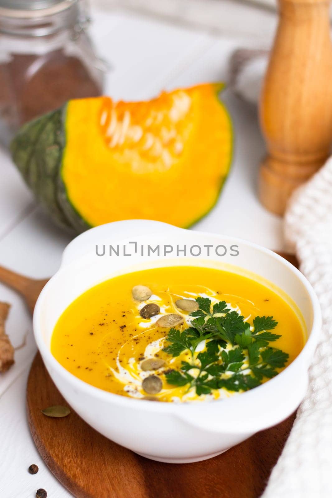 A plate of yellow pumpkin cream soup with cream, herbs and seeds on a white table near ginger, raw pumpkin and a wooden spoon. Vertical