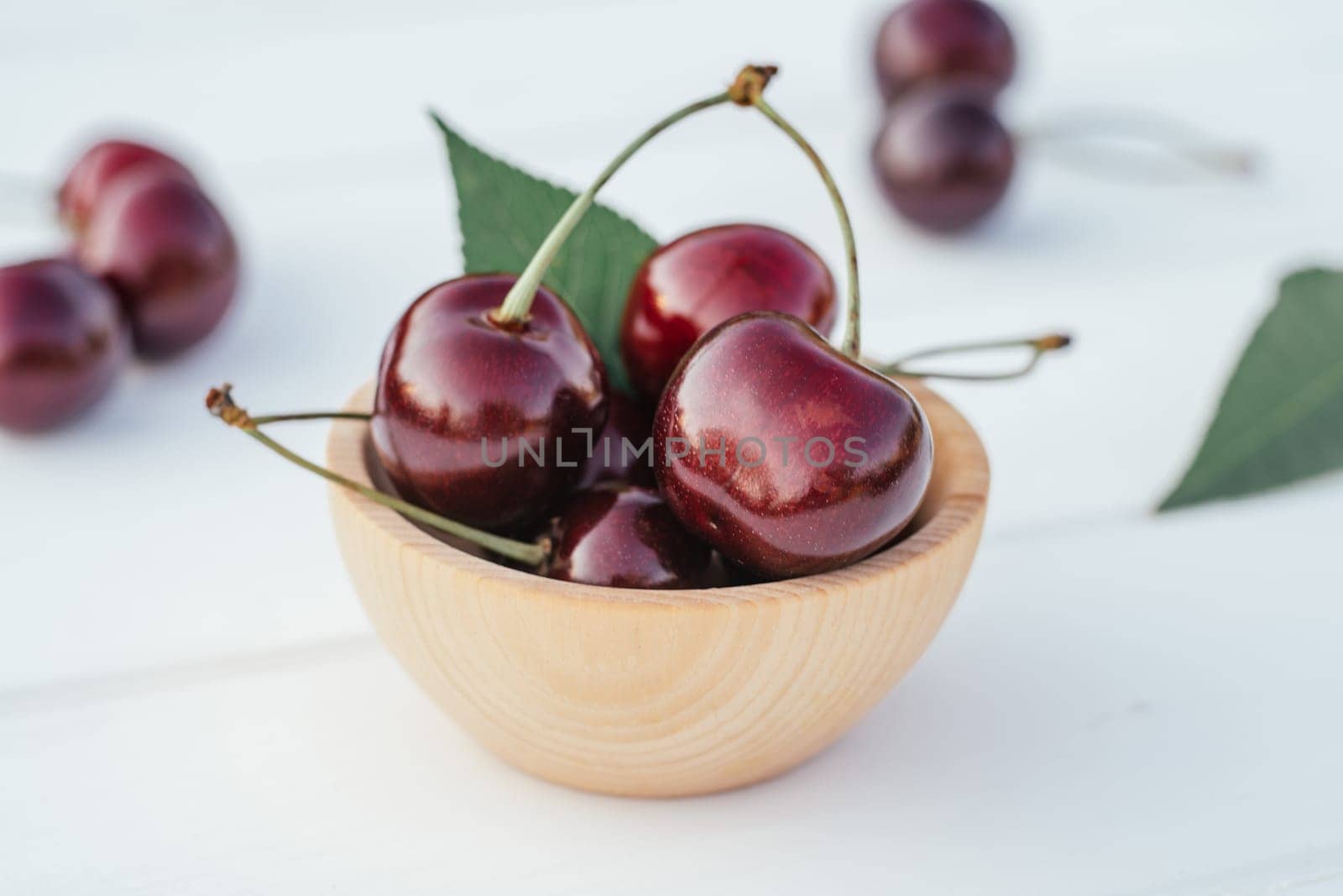 Red ripe sweet cherries berries in bowl standing on wooden white table with green leaves