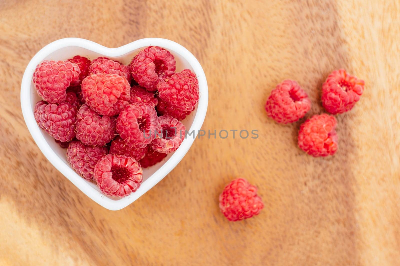 Heart shaped bowl full of ripe red juicy raspberries on wooden background. Top view. Copy space