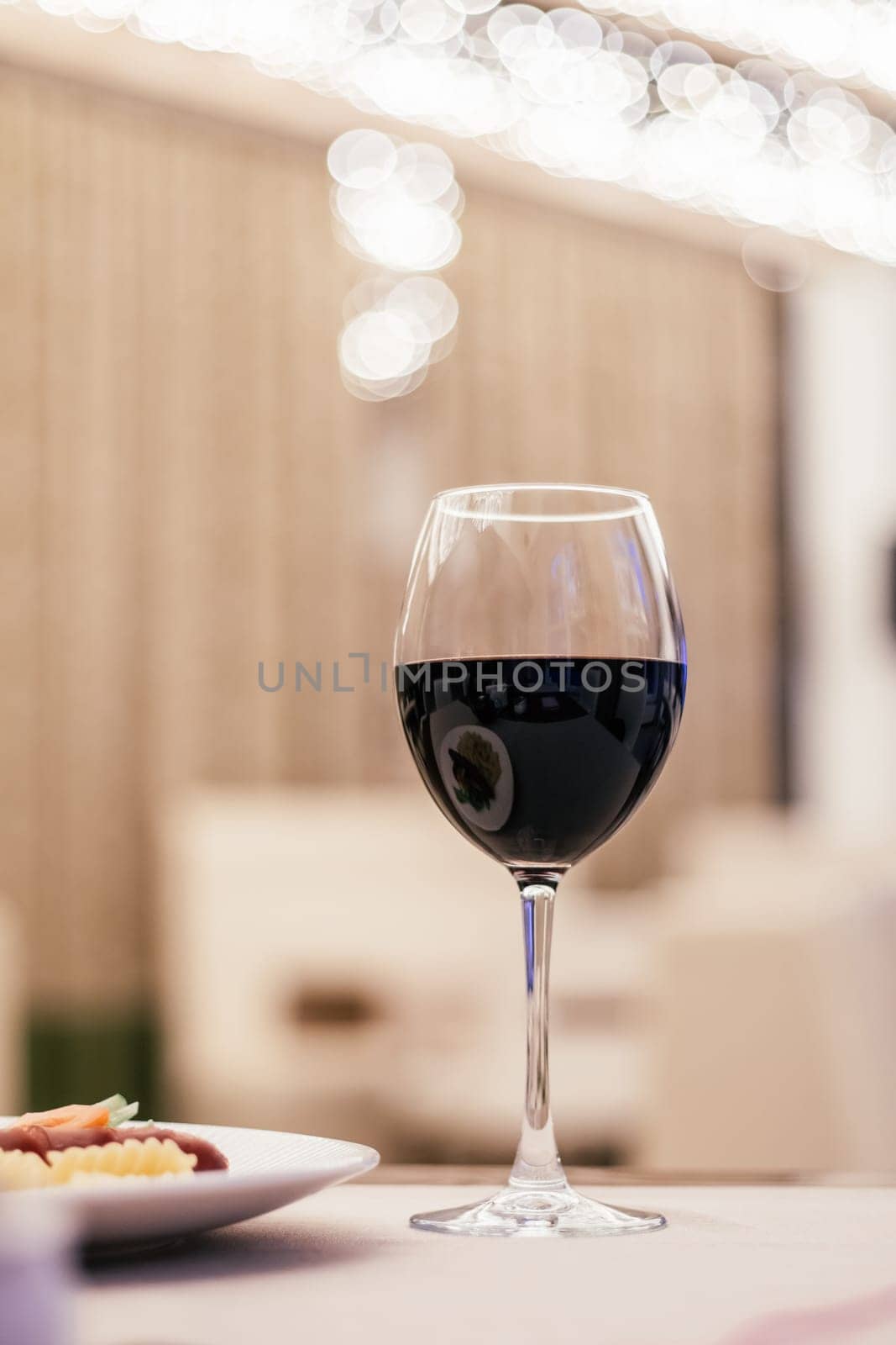 Vertical photo of dark dry wine vine glass on blurred background. Light restaurant, cafe indoor. Plate side dish of fusilli pasta and sausage on white plate on table. Restaurant reservations. Close up