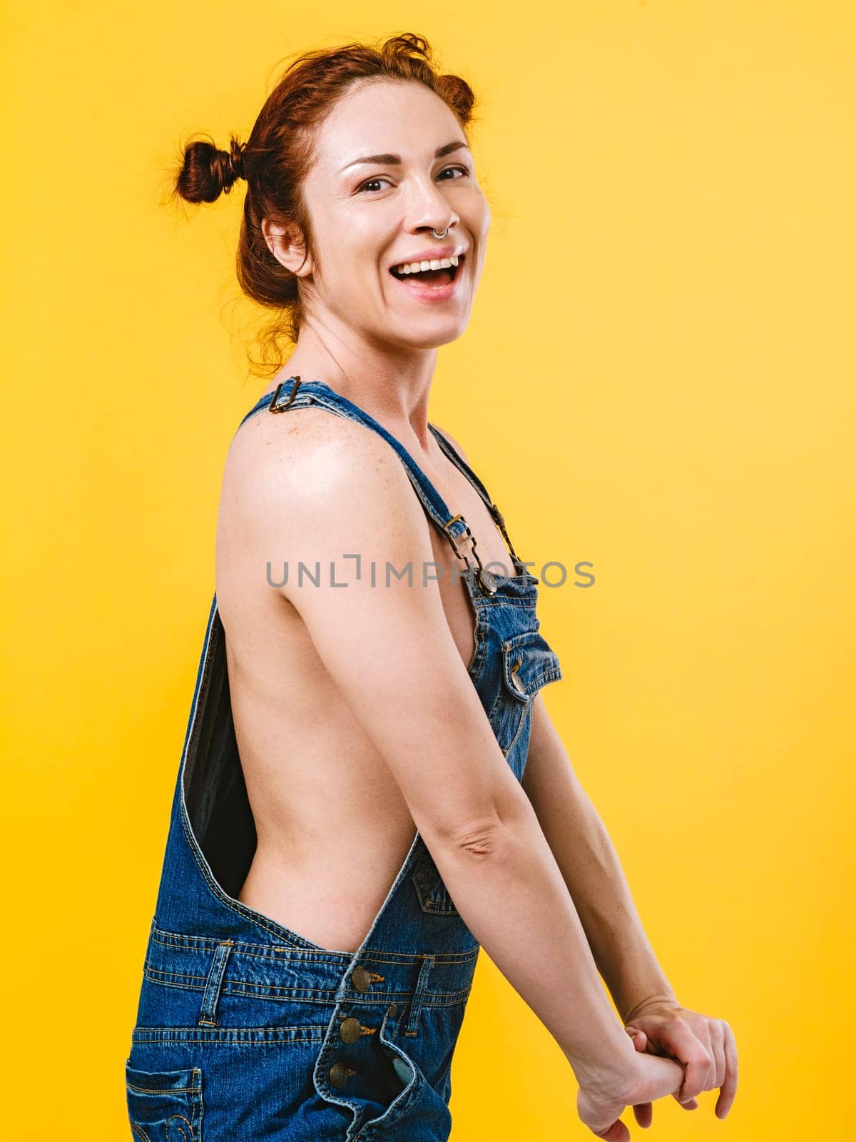 Sexy topless redhead woman laughing and wearing overalls over yellow background.