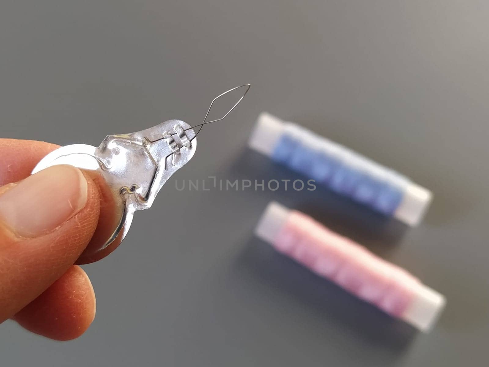 device for drawing the thread into the eye of the needle. Bobbins with blue and pink threads in the background. A woman holds a needle threader in her hand and shows its thin wire eye.