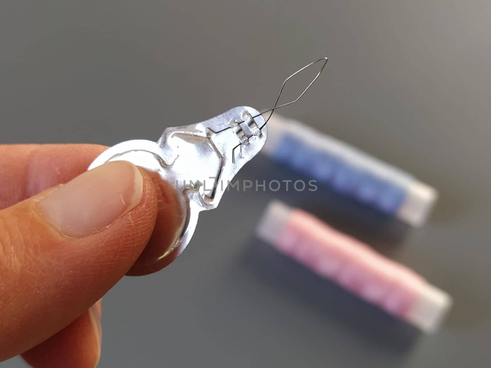device for drawing the thread into the eye of the needle. Bobbins with blue and pink threads in the background. A woman holds a needle threader in her hand and shows its thin wire eye.