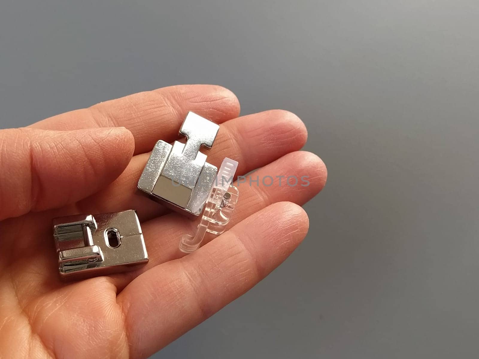 A set of feet for an electric sewing machine in a female hand. Gray backdrop. Standard sewing or zigzag foot, for sewing in zippers, overlock foot and for buttons.
