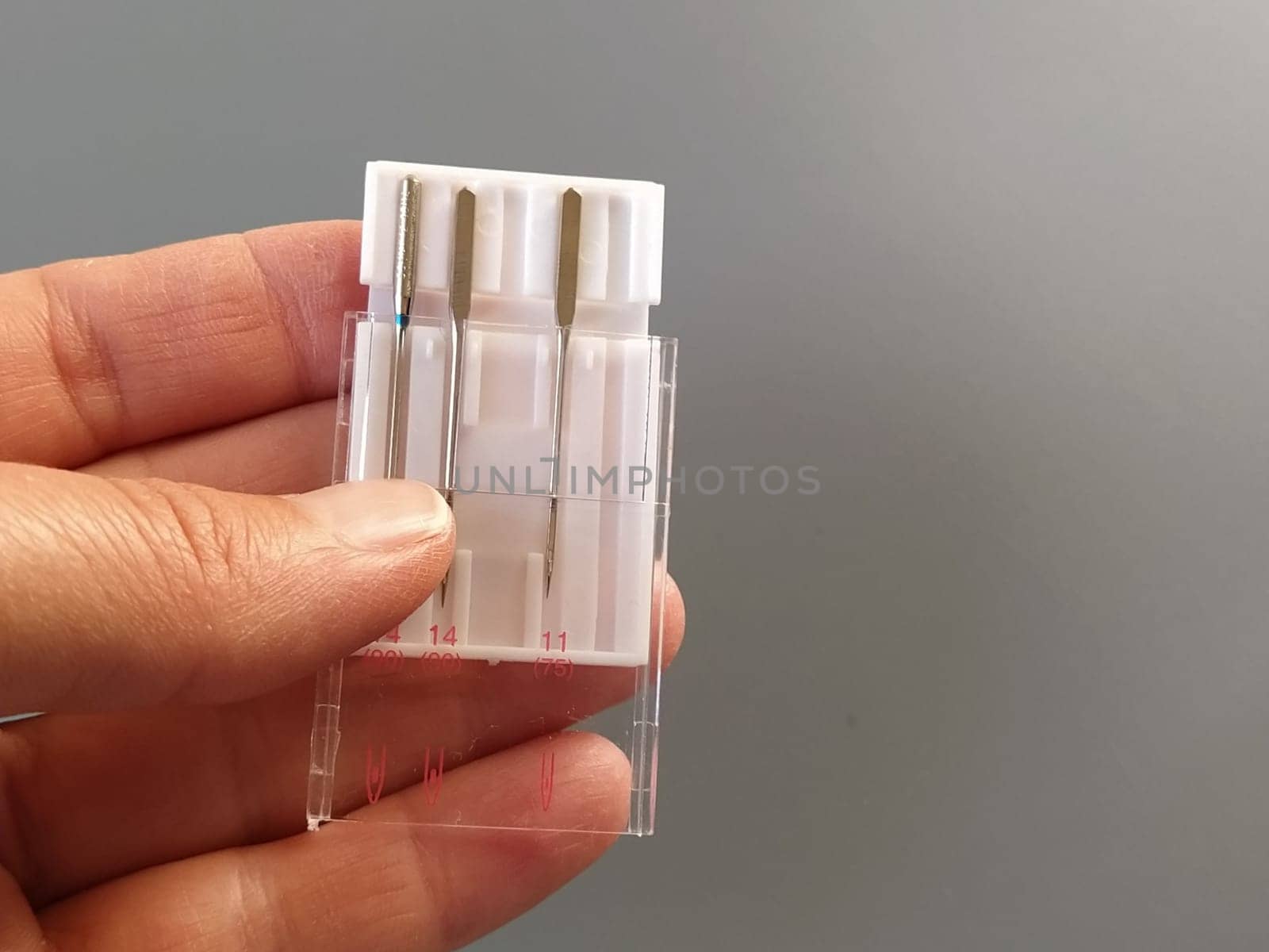a set of sewing machine needles in a white plastic case. A woman's hand holds the needles for an electric sewing machine. Gray background. Different needle thicknesses.