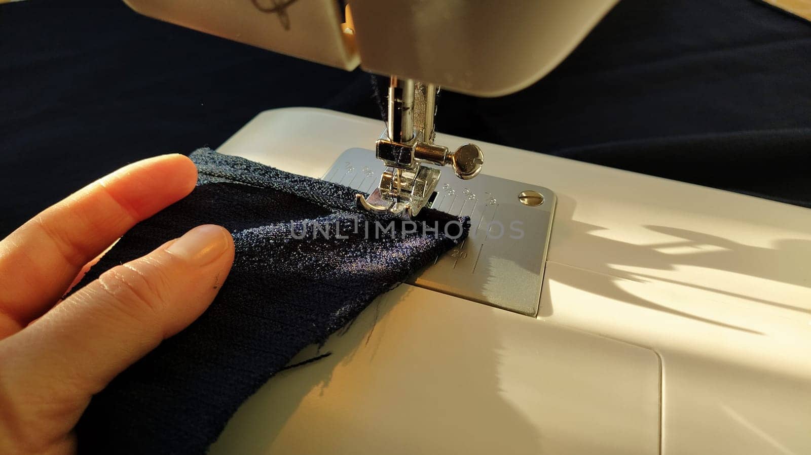 Sewing process on a modern sewing machine, close-up. Sewing machine foot with a needle. Dark blue fabric is trimmed with double wrapping of the fabric edge. Natural sunlight.