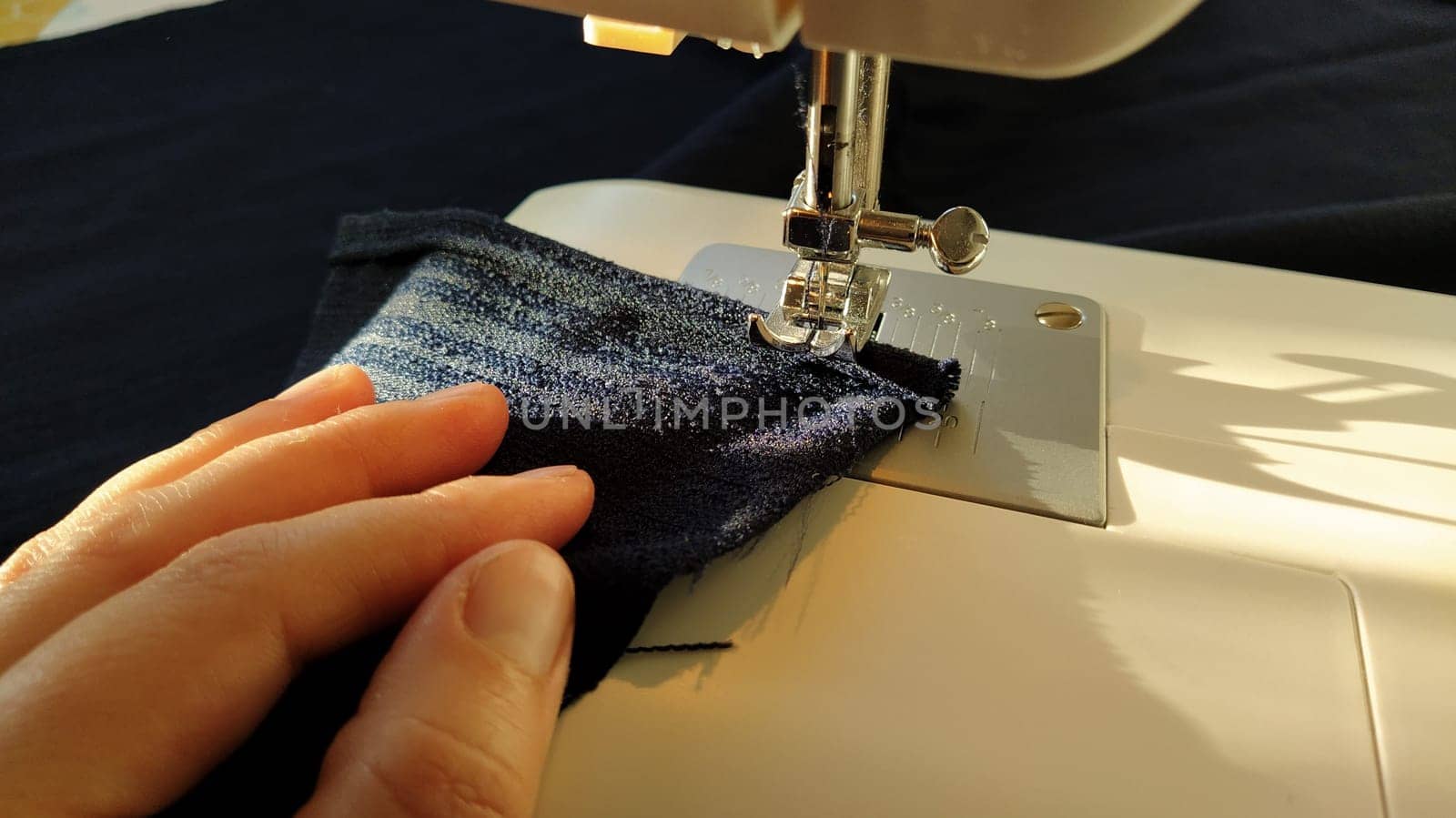 Sewing process on a modern sewing machine, close-up. Sewing machine foot with a needle. Dark blue fabric is trimmed with double wrapping of the fabric edge. Natural sunlight.