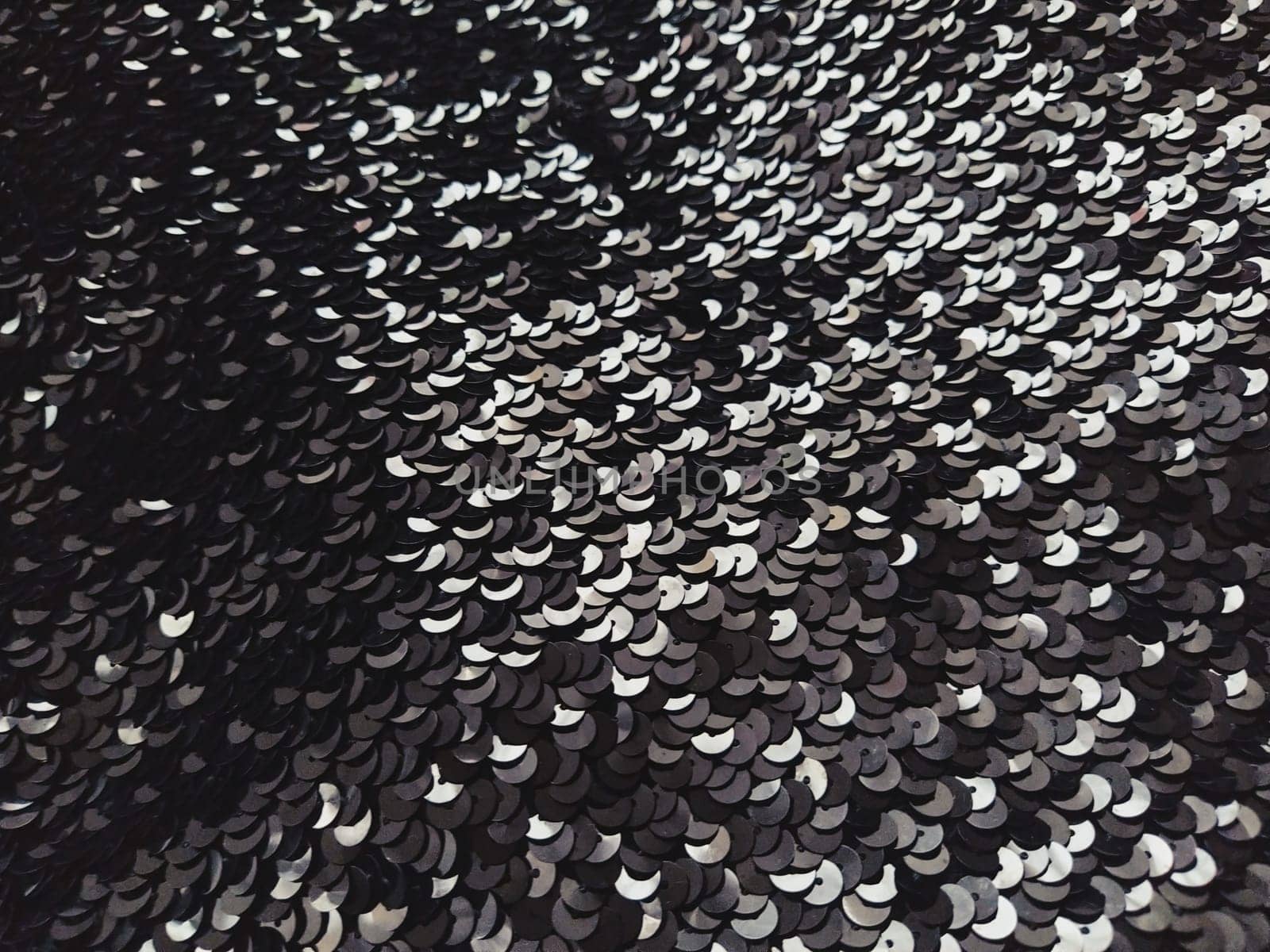 Sequin fabric. Black sequins with white and gray sheen. Reflection of light on individual elements in a chaotic manner. Sewing or Christmas decorative background.