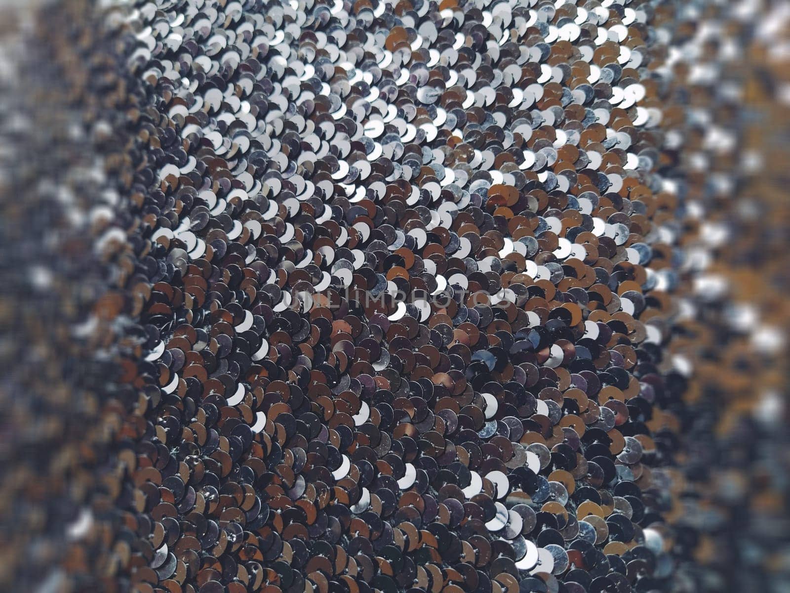 Sequin fabric. Black sequins with white-gray tint. Reflection of light on individual elements in a chaotic manner. Background blurred on the left and right. Soft focus.