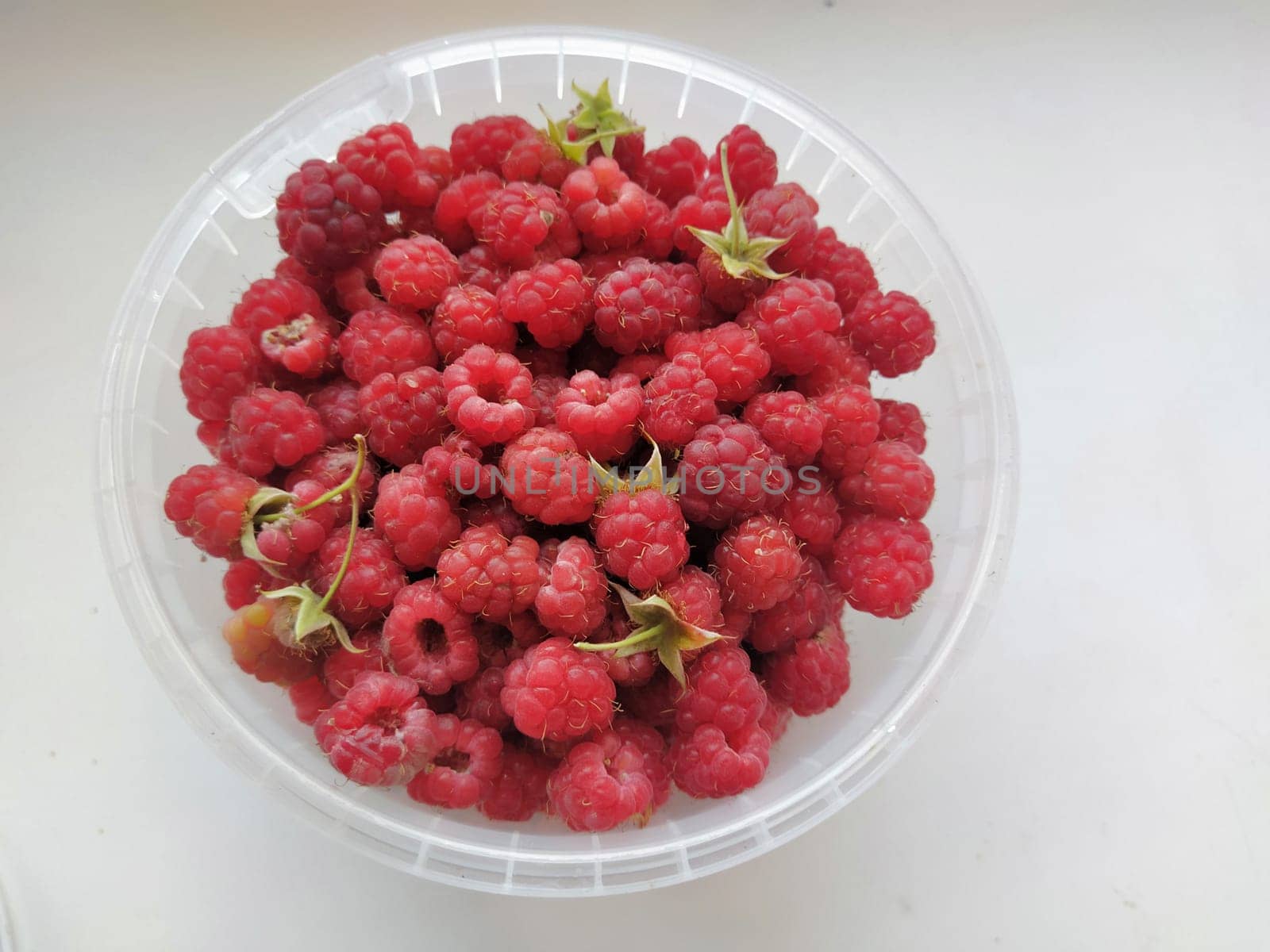 Red juicy raspberries in plastic box container on white background, top view. Raspberries in a plastic bucket. Mellow razz
