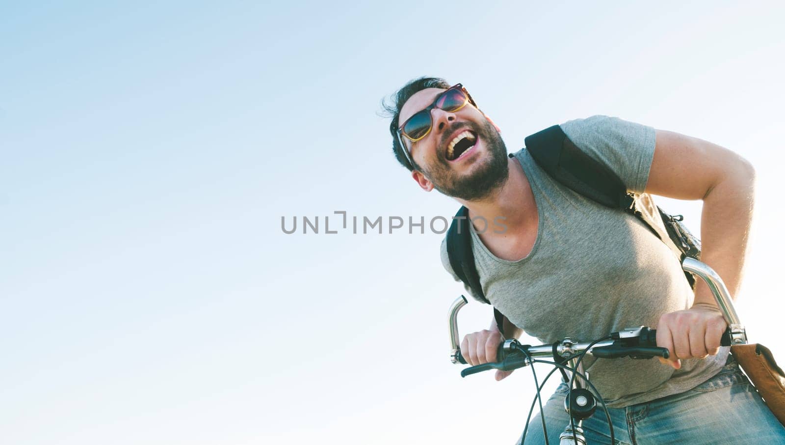 Enthusiastic hipster guy with retro sunglasses having outdoor fun and riding a bicycle. Active sport man with excited face expression exploring and traveling by mountain bike on the road. Vintage film filter style image.