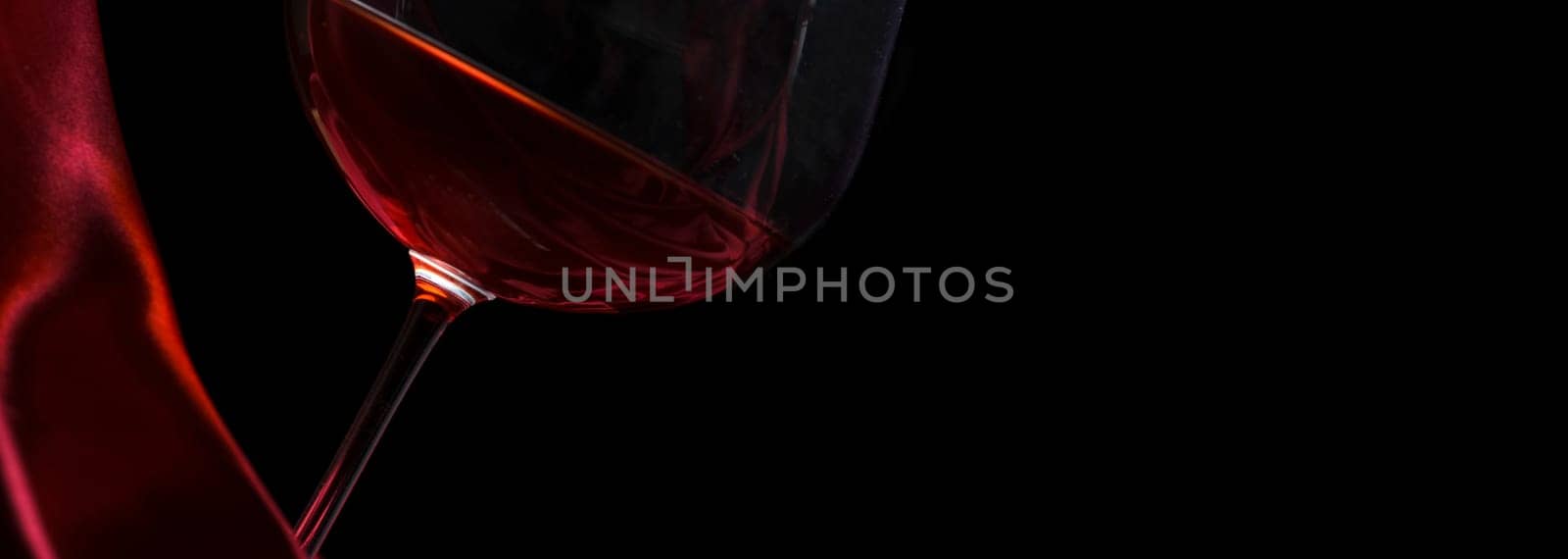 Glass of red wine on red silk against black background. Wine list design layout. Luxury restaurant romantic dinner concept. Closeup wineglass on dark backdrop.