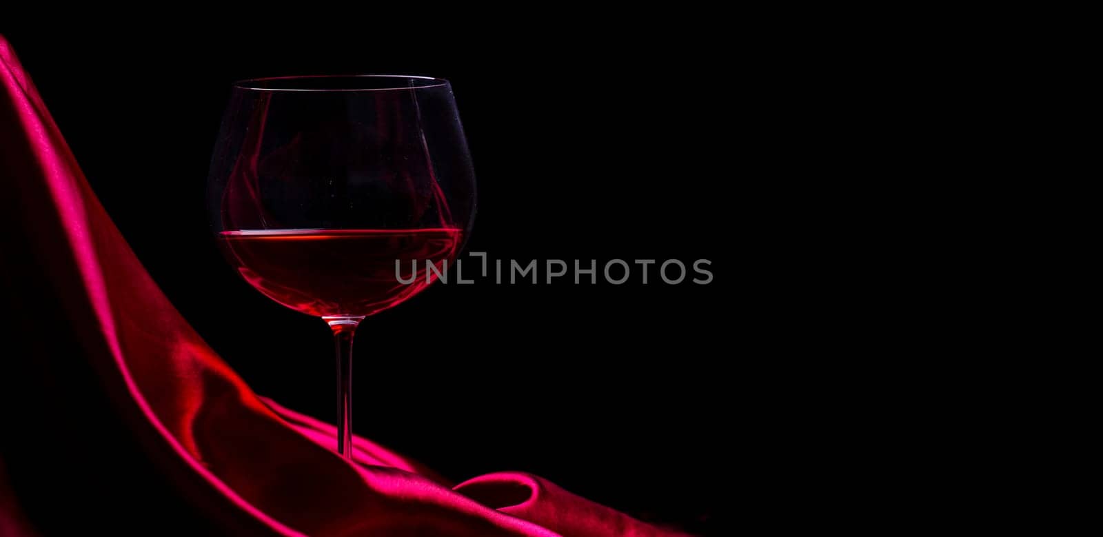 Glass of red wine on red silk against black background. Wine list design background. Luxury restaurant romantic dinner concept.