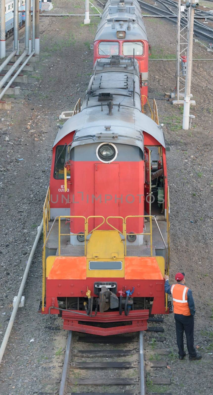 Shunting locomotive standing on rails at the train station. Nearby stands a railway worker in overalls. Front view from the top.