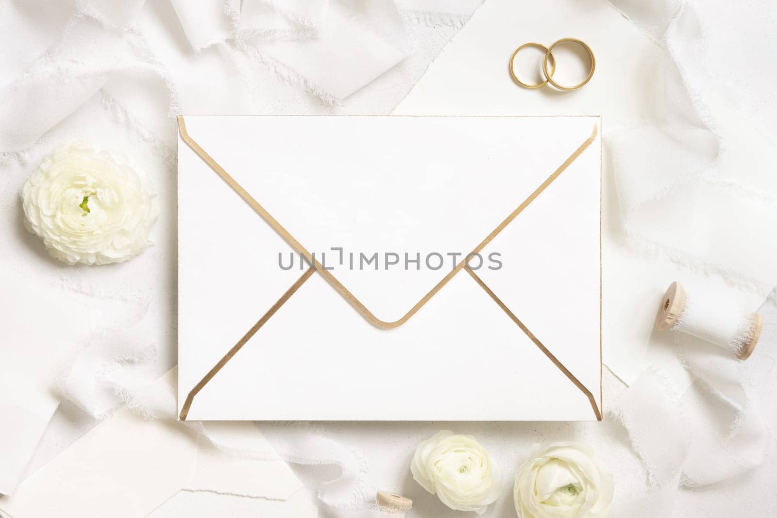 Blank envelope near cream roses, white silk ribbons and wedding rings top view, wedding mockup. Romantic scene with vertical card and pastel flowers flat lay. Valentines, Spring or Mothers day concept