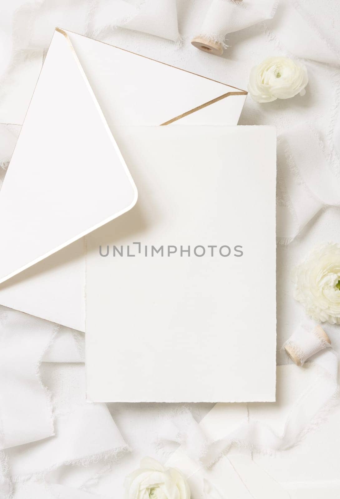Blank card and envelope near cream roses and white silk ribbons top view, wedding mockup. Romantic scene with vertical card and pastel flowers flat lay. Valentines, Spring or Mothers day concept
