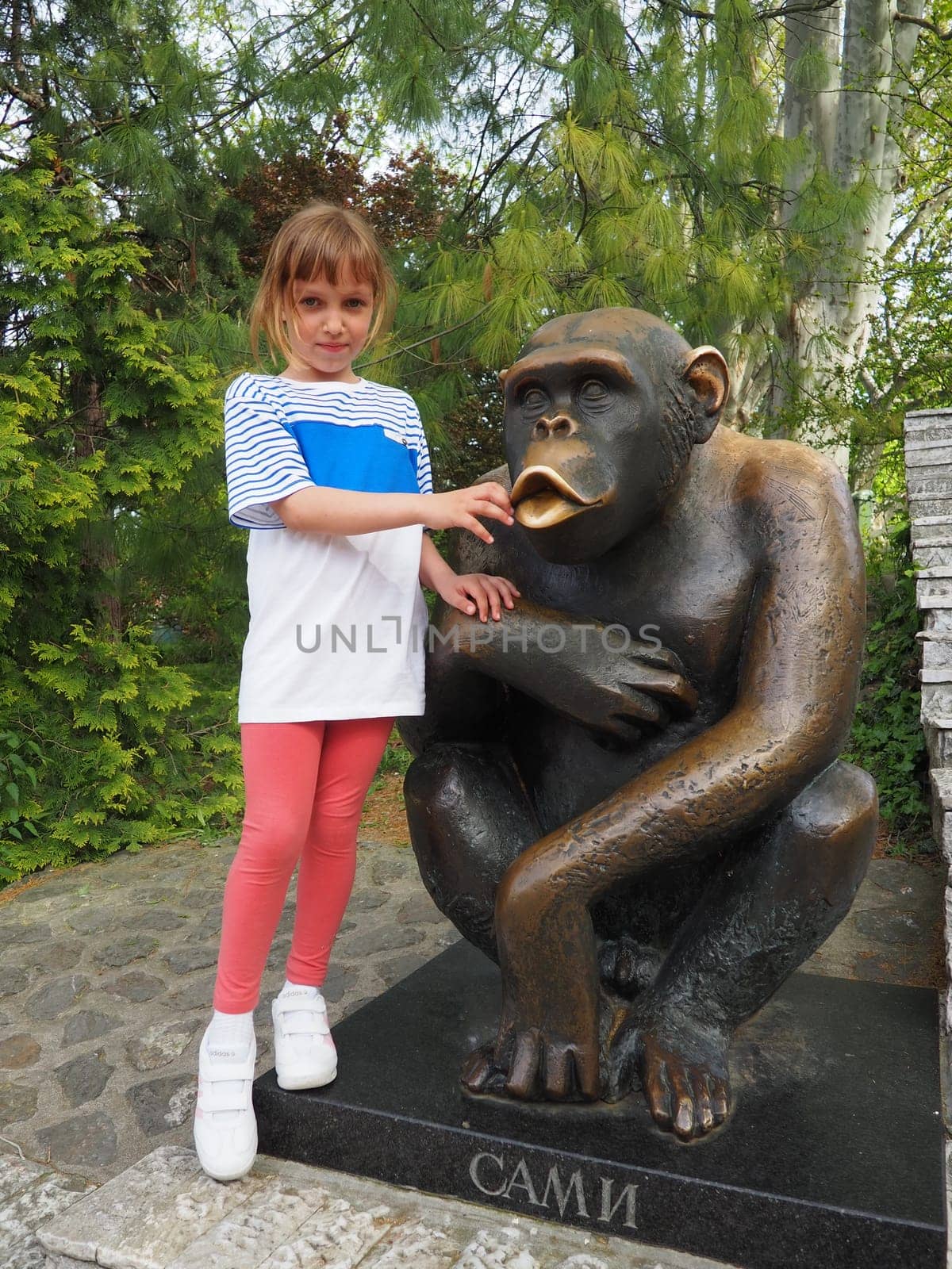 Belgrade, Serbia, April 25, 2021. A 7-year-old girl with blond hair stands next to the Sami chimpanzee statue, the symbol of the Belgrade Zoo. Inscription - Sami, monkey's nickname