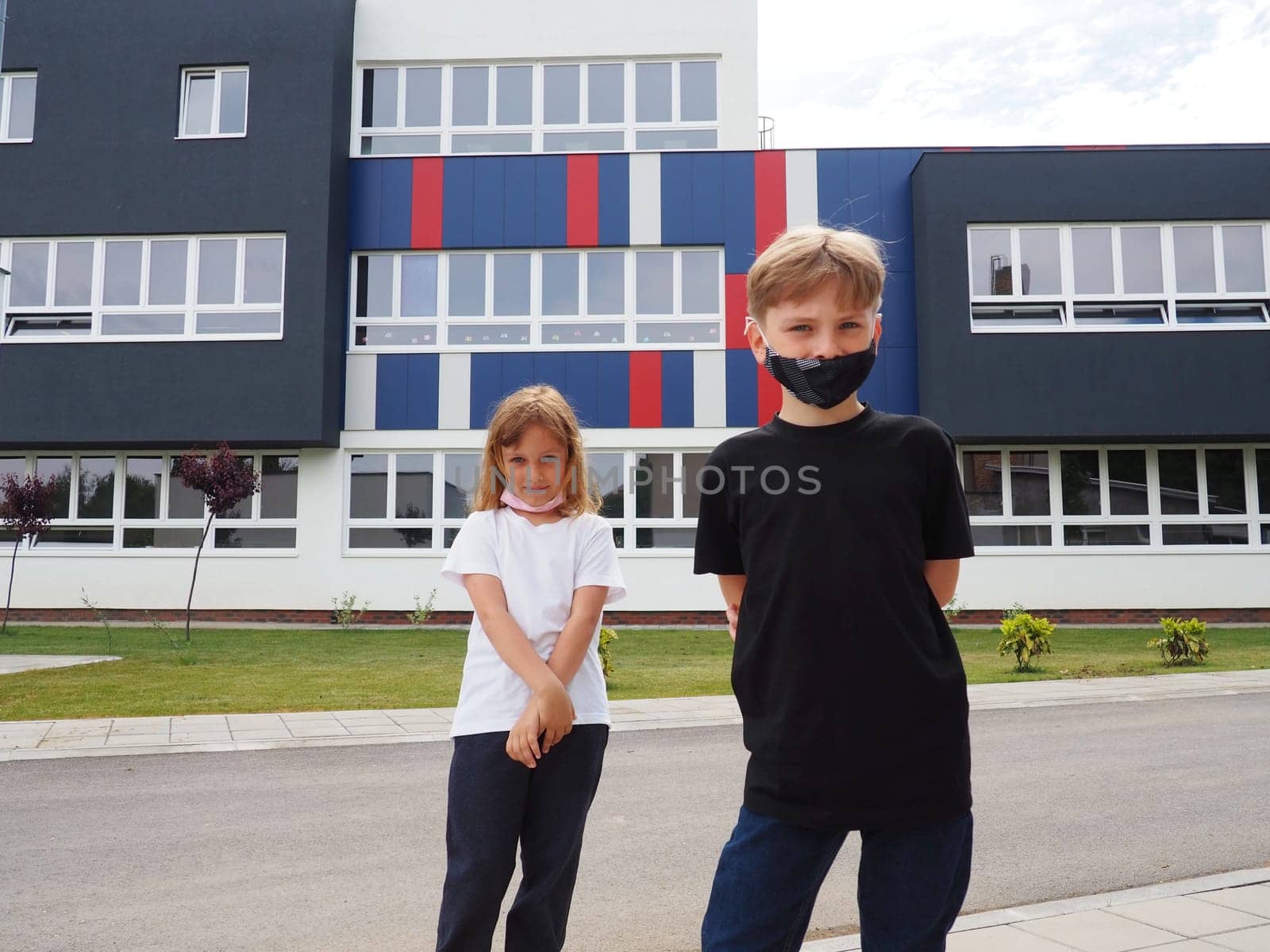 A blond boy 8 years old and a blond girl 7 years old are standing against the background of a modern school and looking at the camera. Children wear face masks. Black and white t-shirts.