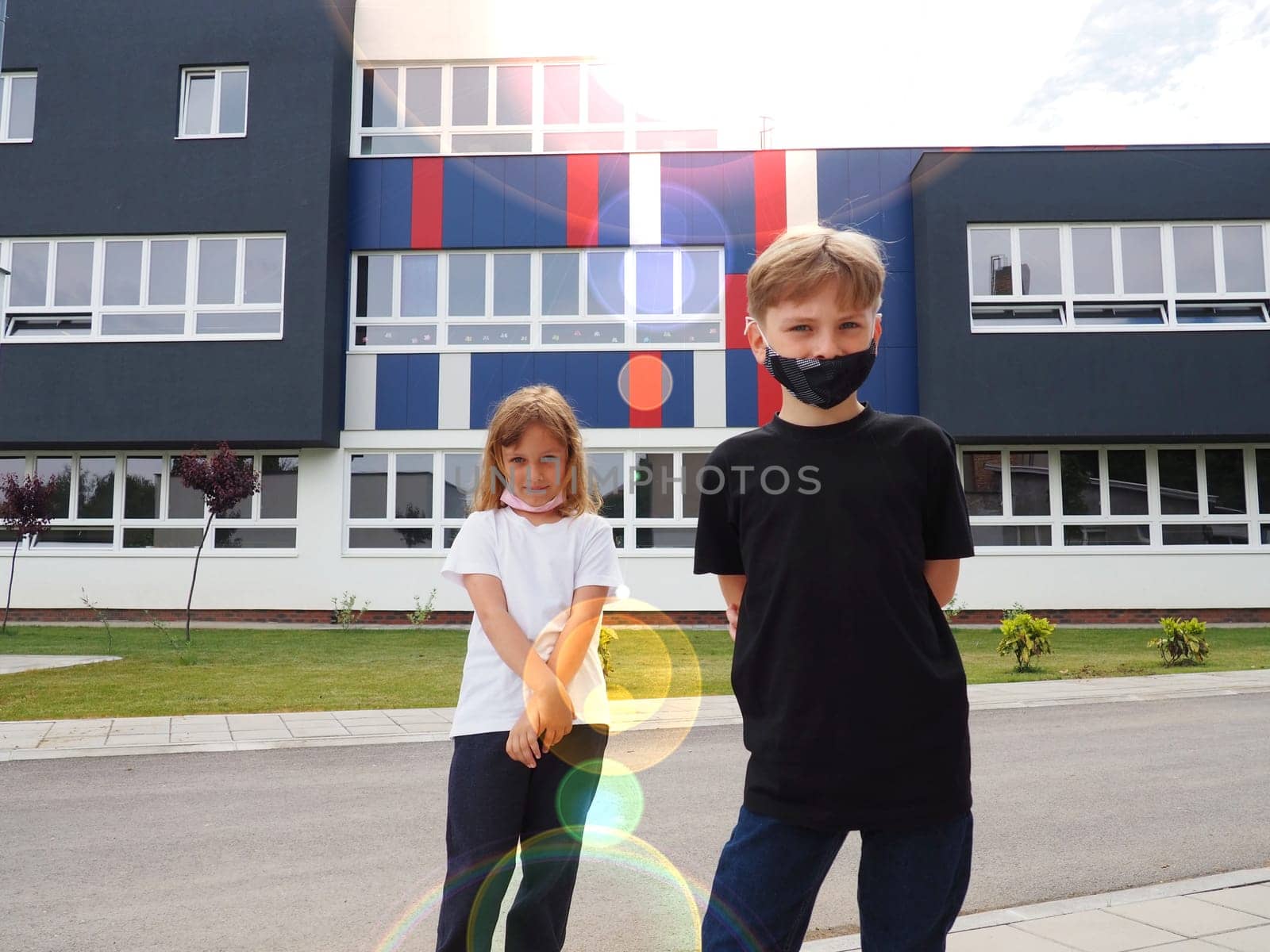 A blond boy 8 years old and a blond girl 7 years old are standing against the background of a modern school and looking at the camera. Children wear face masks. Sun glare.