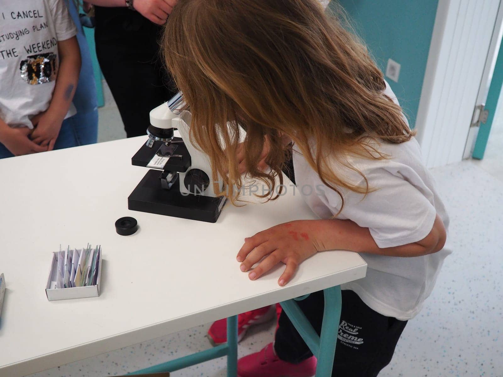 Sremska Mitrovica, Serbia, May 15, 2021. A girl of 7 years old in a white T-shirt looks with one eye through a microscope. Biology lesson. The process of teaching children. School lessons.