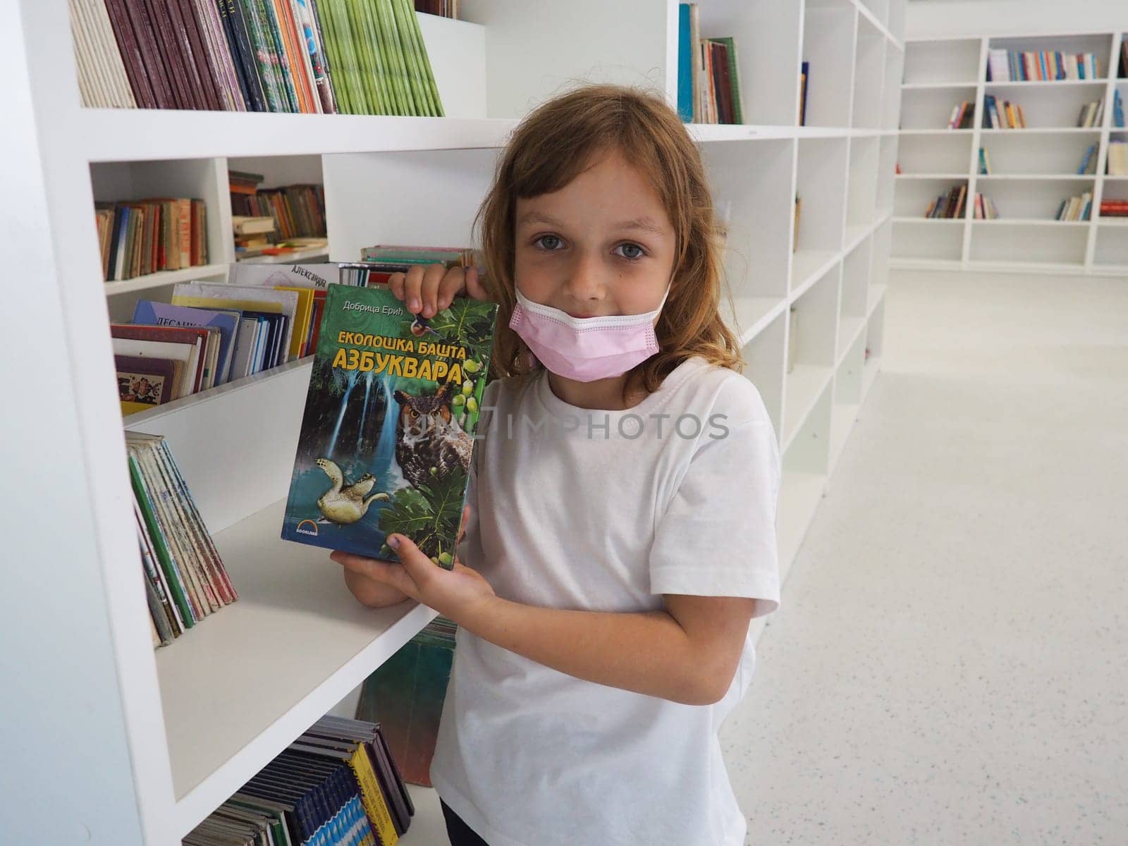 Sremska Mitrovica, Serbia May 15, 2021 A 7-year-old girl wearing a mask and a white T-shirt stands near a bookshelf in a library and touches books with hands. School education, literature and reading.