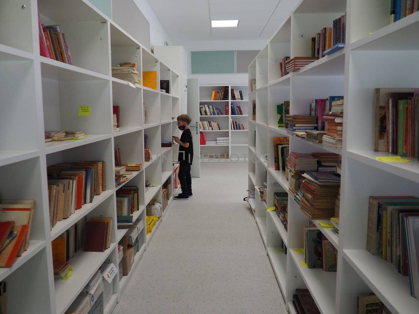 Sremska Mitrovica, Serbia May 15 2021 An 8-year-old boy wearing a mask and a black T-shirt stands near the bookshelf in the library and reads a book. School education. Love for literature and reading.