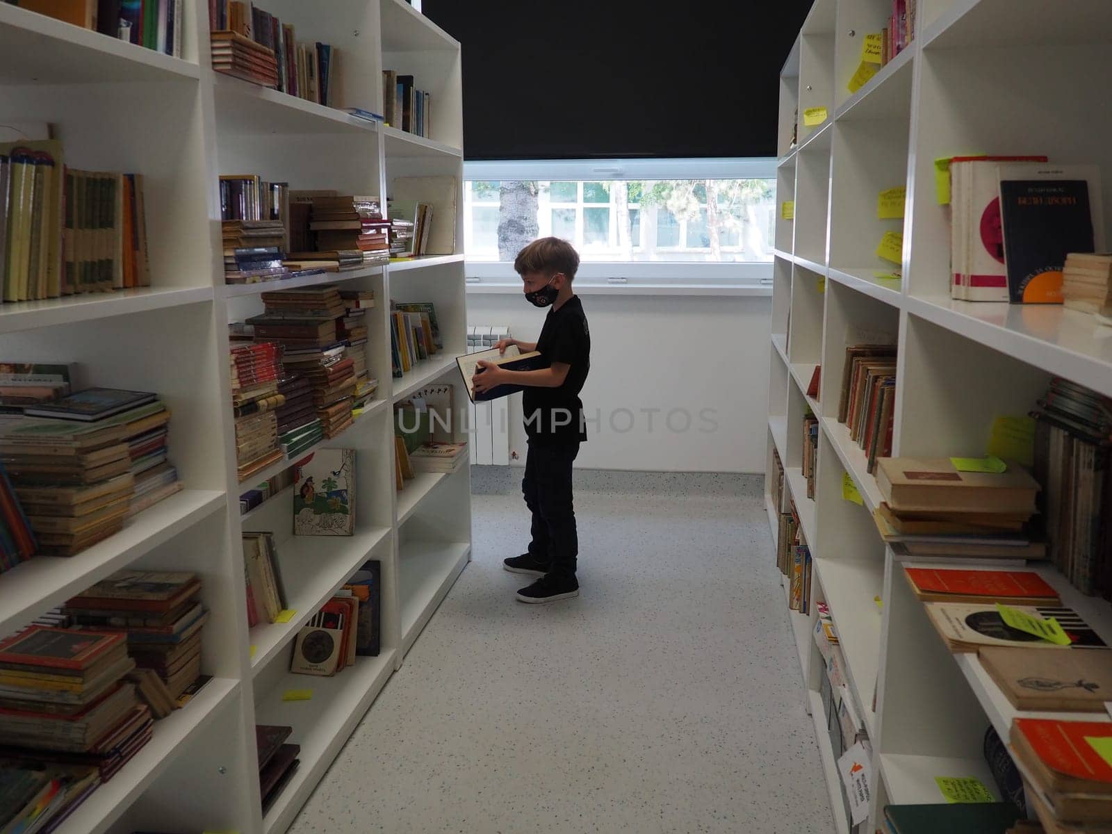 Sremska Mitrovica, Serbia May 15 2021 An 8-year-old boy wearing a mask and a black T-shirt stands near the bookshelf in the library and reads a book. School education. Love for literature and reading