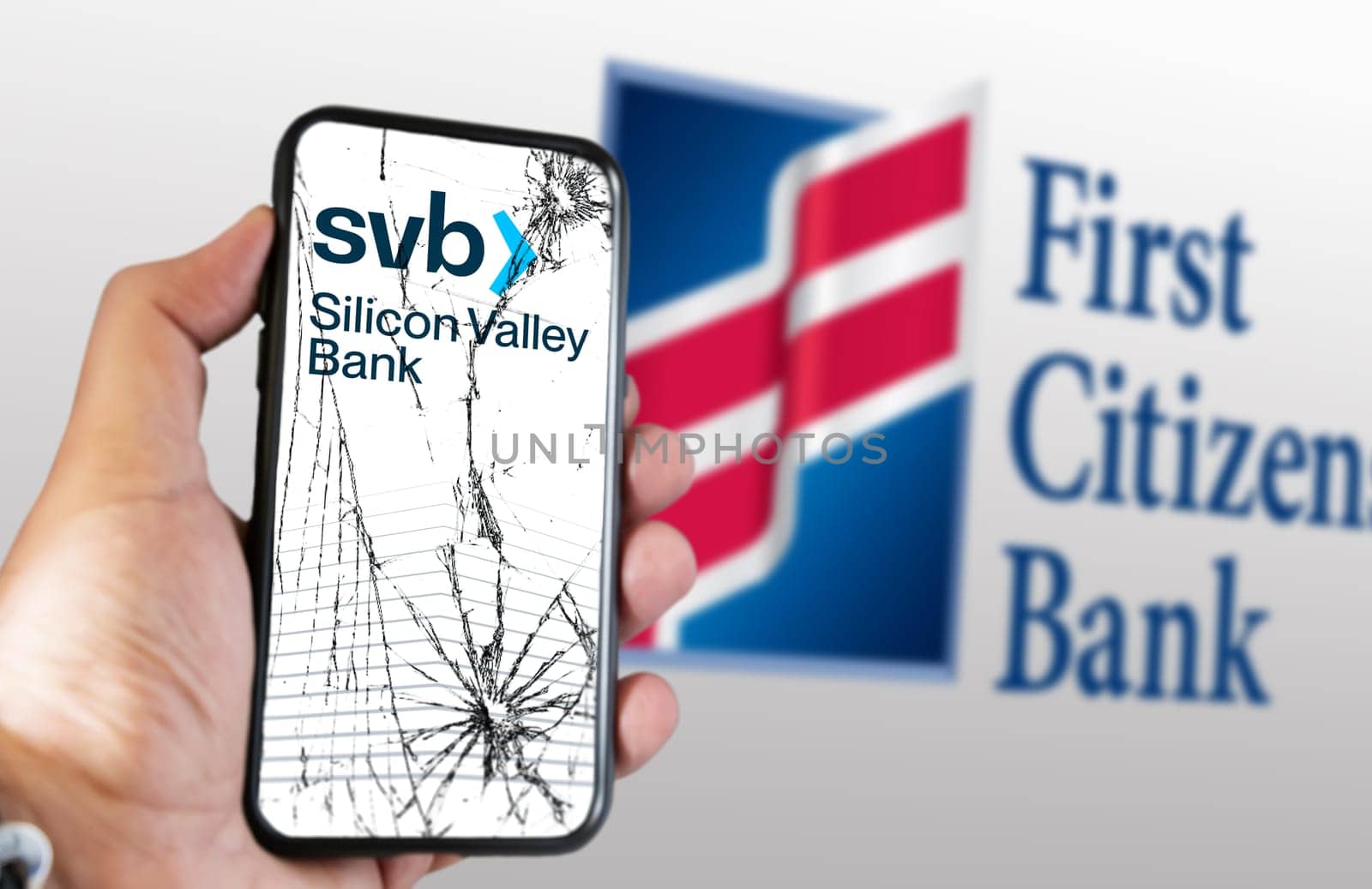 New York, US, March 2023: Hand holding a phone with Silicon Valley Bank logo on cracked screen. First Citizens Bank logo blurred on white background. Illustrative editorial