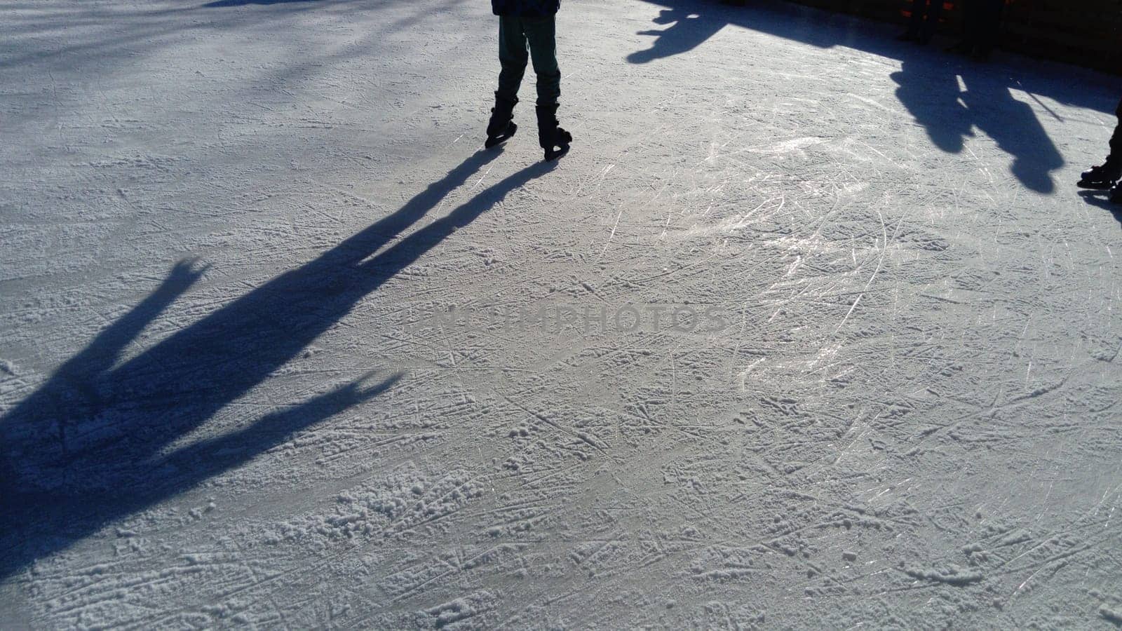 Children ride in a city park on an ice rink. Feet skater while skating on ice. The low winter sun weakly illuminates the ice. Dark shapes and long shadows on the surface. Sports movements.