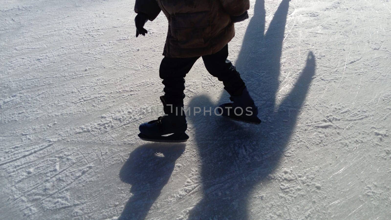 Children ride in a city park on an ice rink. Feet skater while skating on ice. The low winter sun weakly illuminates the ice. Dark shapes and long shadows on the surface. Sports movements.